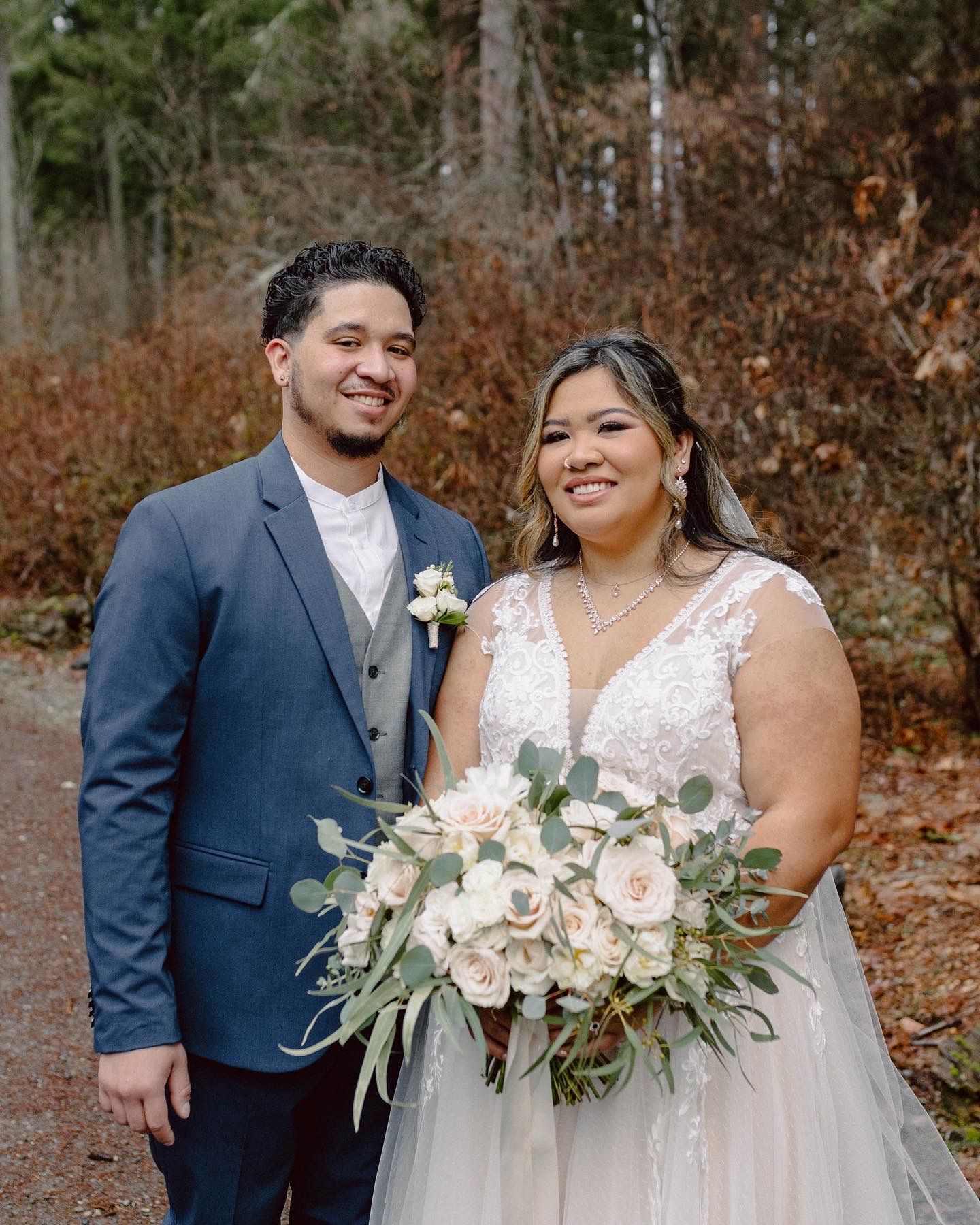 Wedding couple poses with elegant white and green floral bouquets in a forest setting.