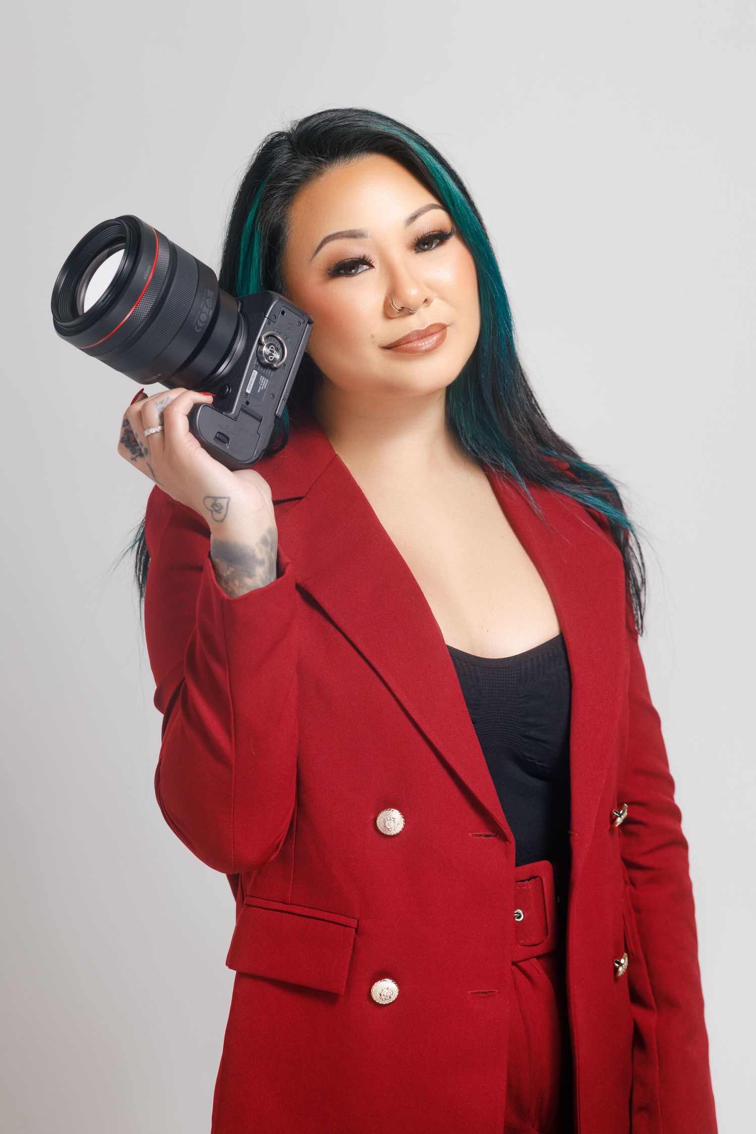 Professional photographer in a red blazer holding a camera against a light gray studio background.