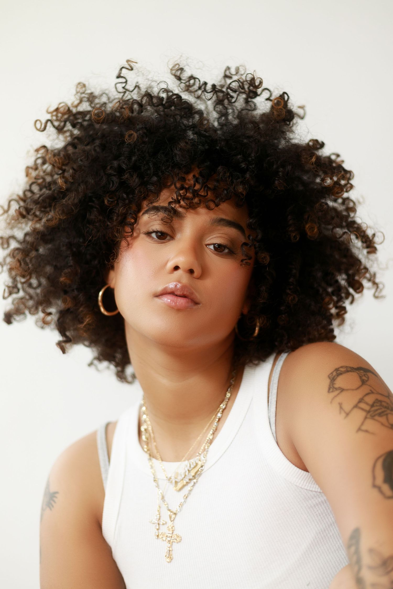 Portrait of person with voluminous curly hair wearing gold necklaces and white tank top.