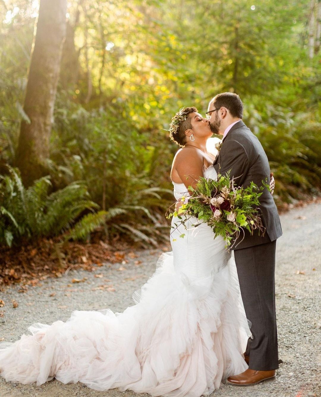 A romantic wedding couple shares a kiss on a sunlit path surrounded by lush greenery.