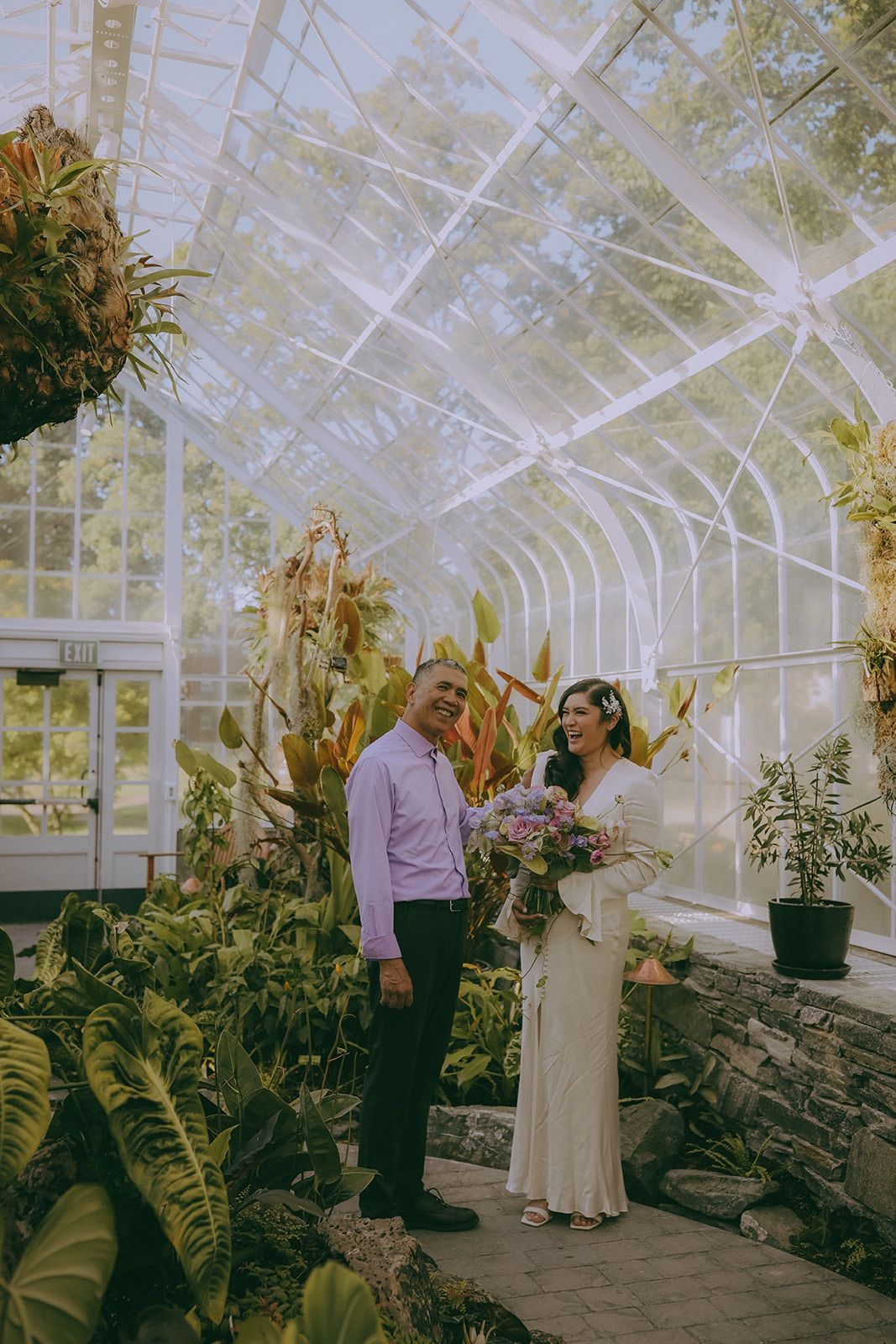 Couple standing together in a greenhouse filled with tropical plants and natural light.