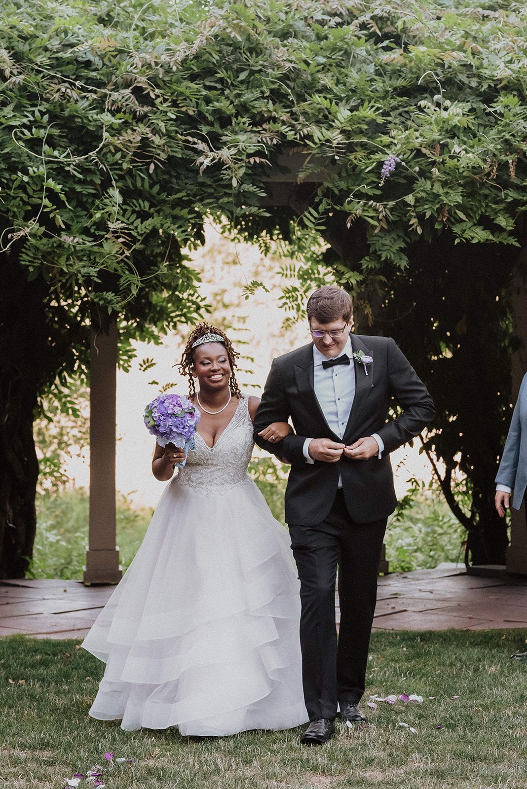 Wedding party members walk and laugh together on a garden path surrounded by greenery.