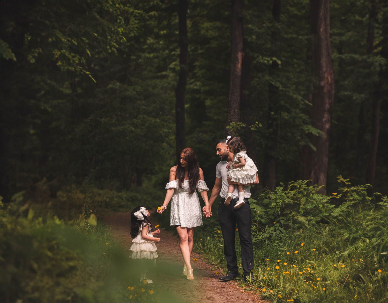 Familj i ljusa sommarkläder promenerar i skogen under en varm sommareftermiddag – naturlig familjefotografering utomhus.