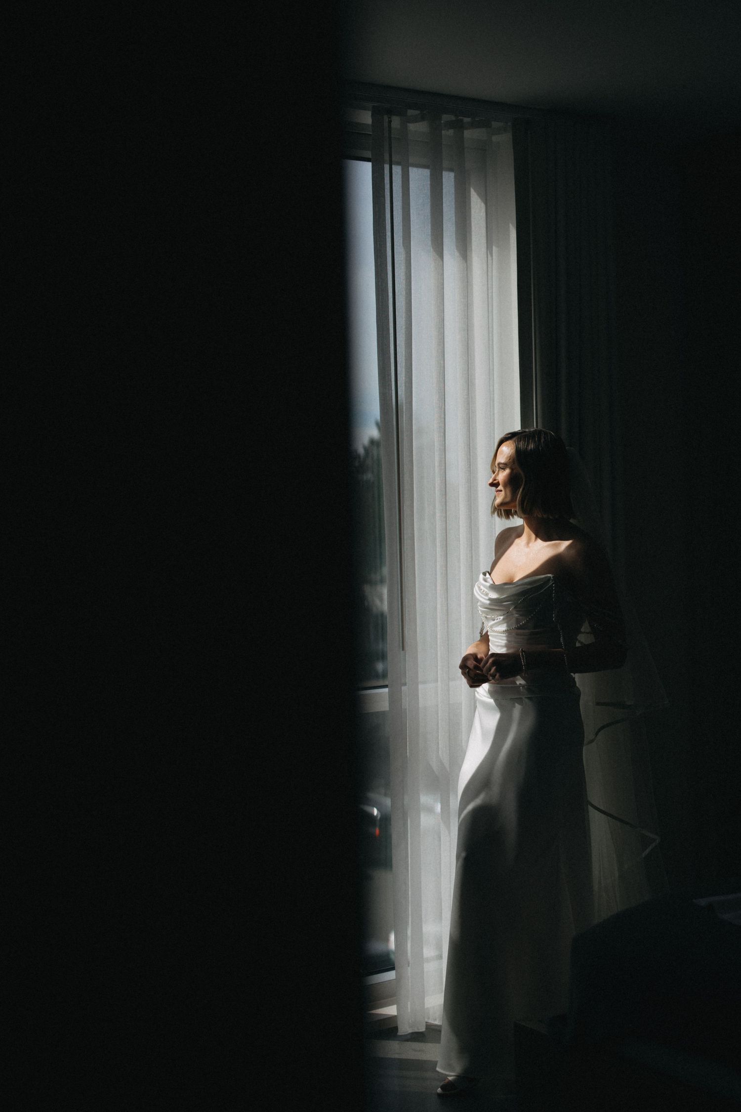 Radiant bride in white strapless gown standing by sunlit window with sheer curtains on wedding day.