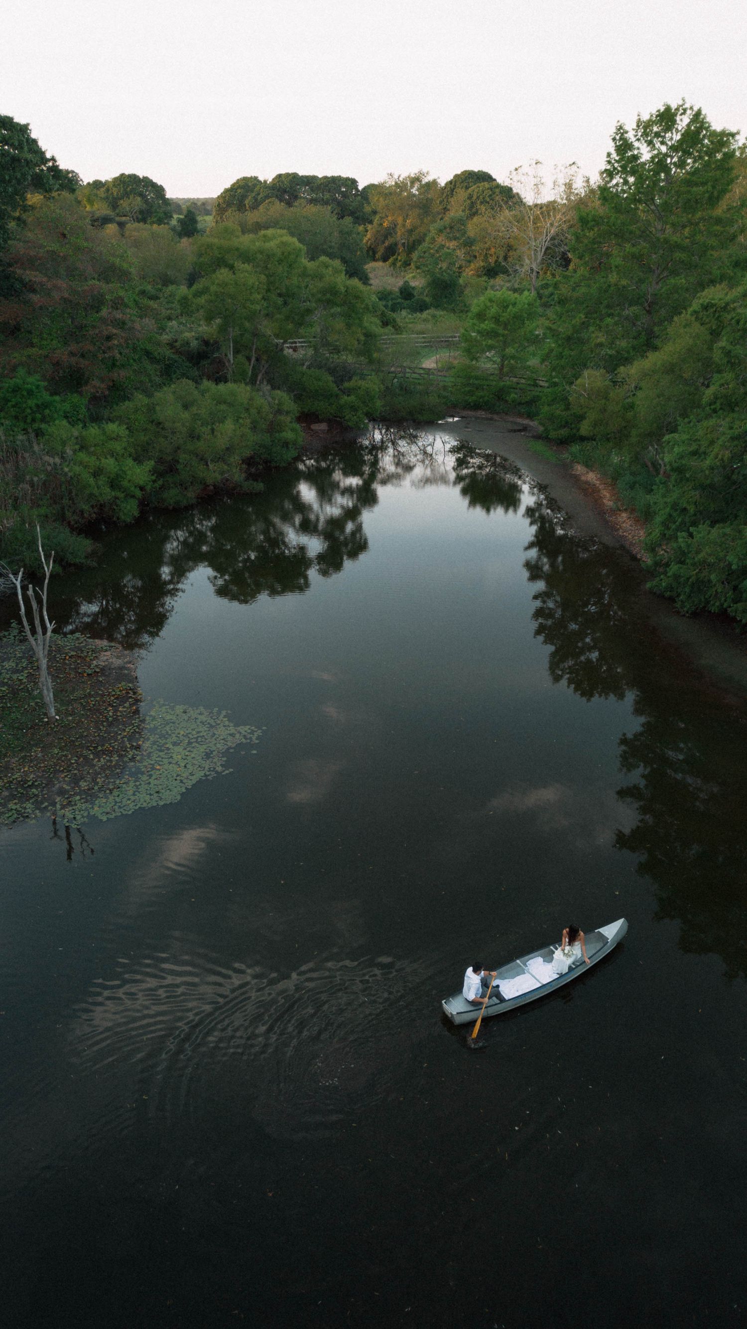 Aerial view of a couple in a white boat gliding along a calm, tree-lined river at dusk.