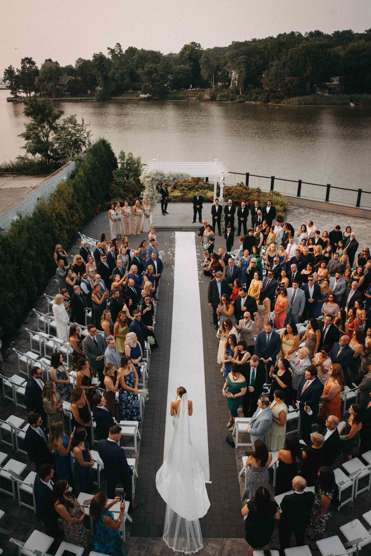 Aerial view of outdoor waterfront wedding ceremony with bride walking white aisle runner toward the altar.