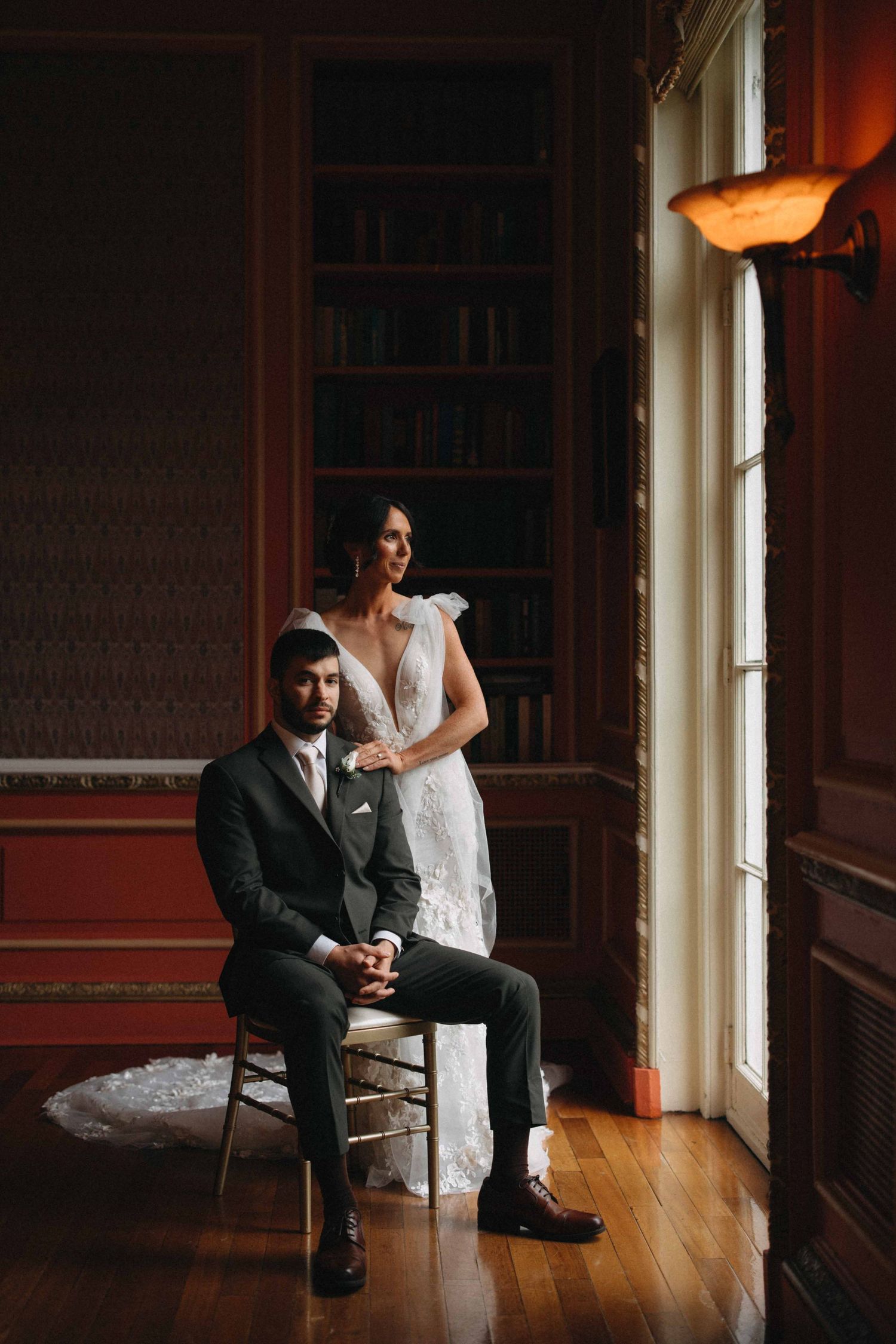 Bride standing beside seated groom in a dark elegant library room with warm window light.