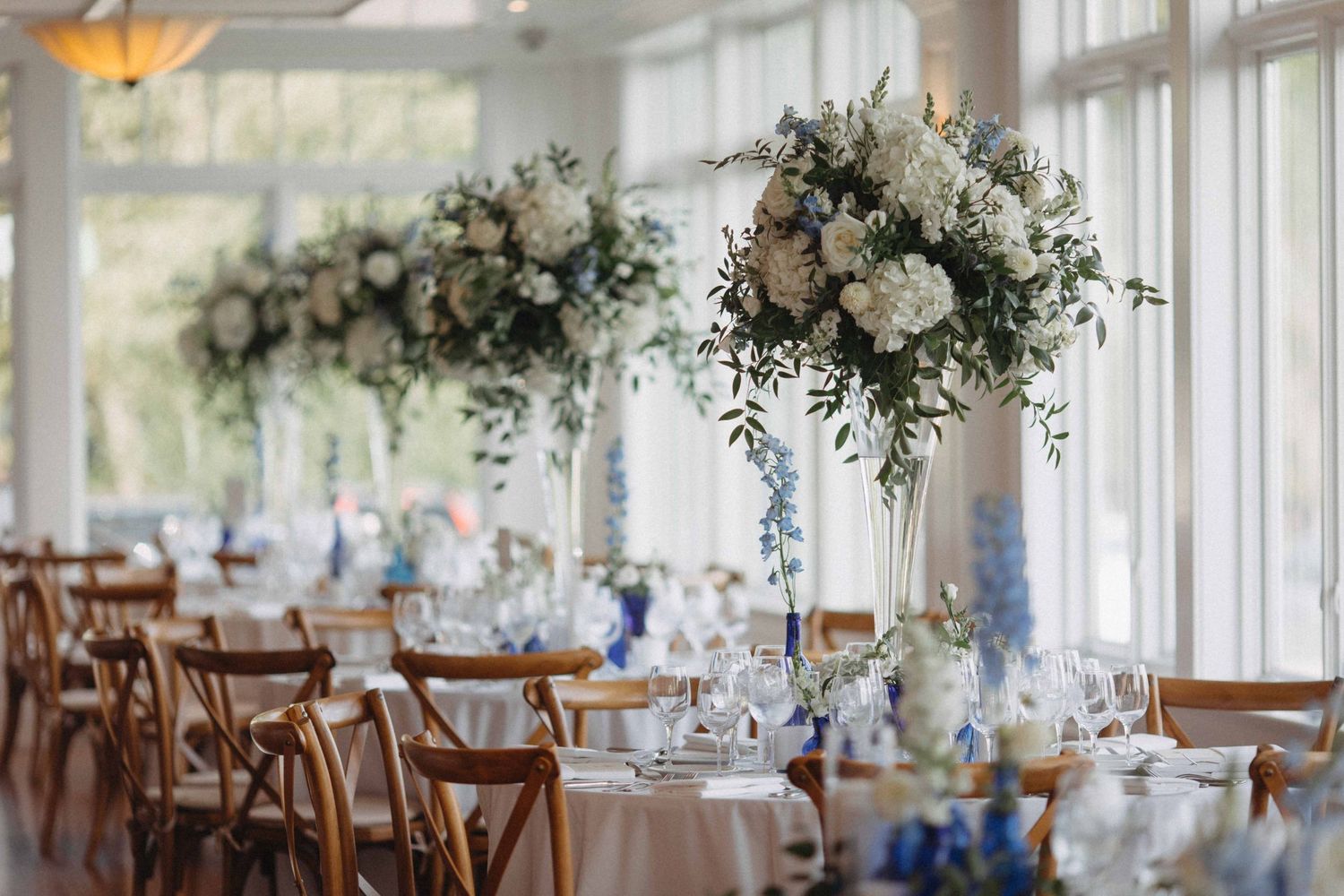 Elegant wedding reception table with tall white floral centerpieces and blue accents in sunlit venue.