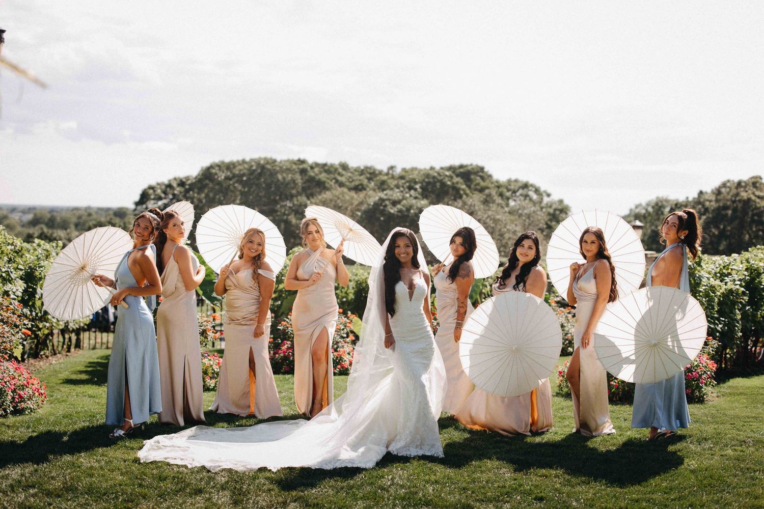 Bride with bridesmaids in pastel dresses holding white parasols in a lush vineyard garden setting.