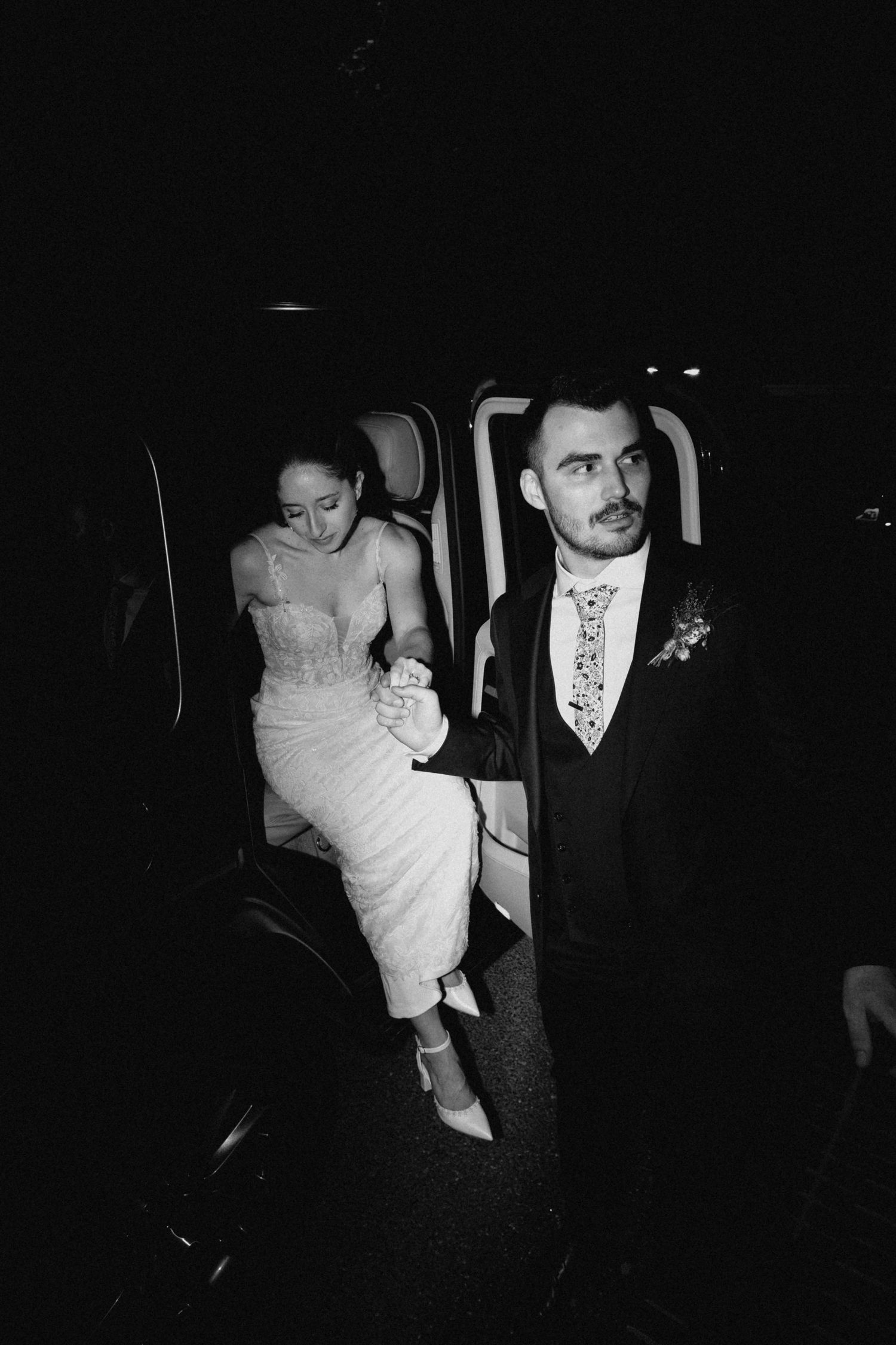 Black and white photo of groom helping bride exit vehicle at night wedding.