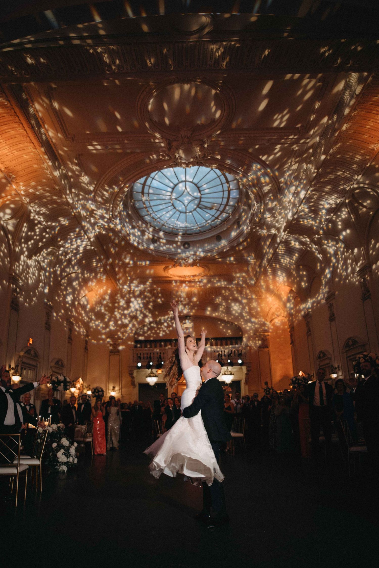 Couple sharing first dance under sparkling lights in grand ballroom with skylight.
