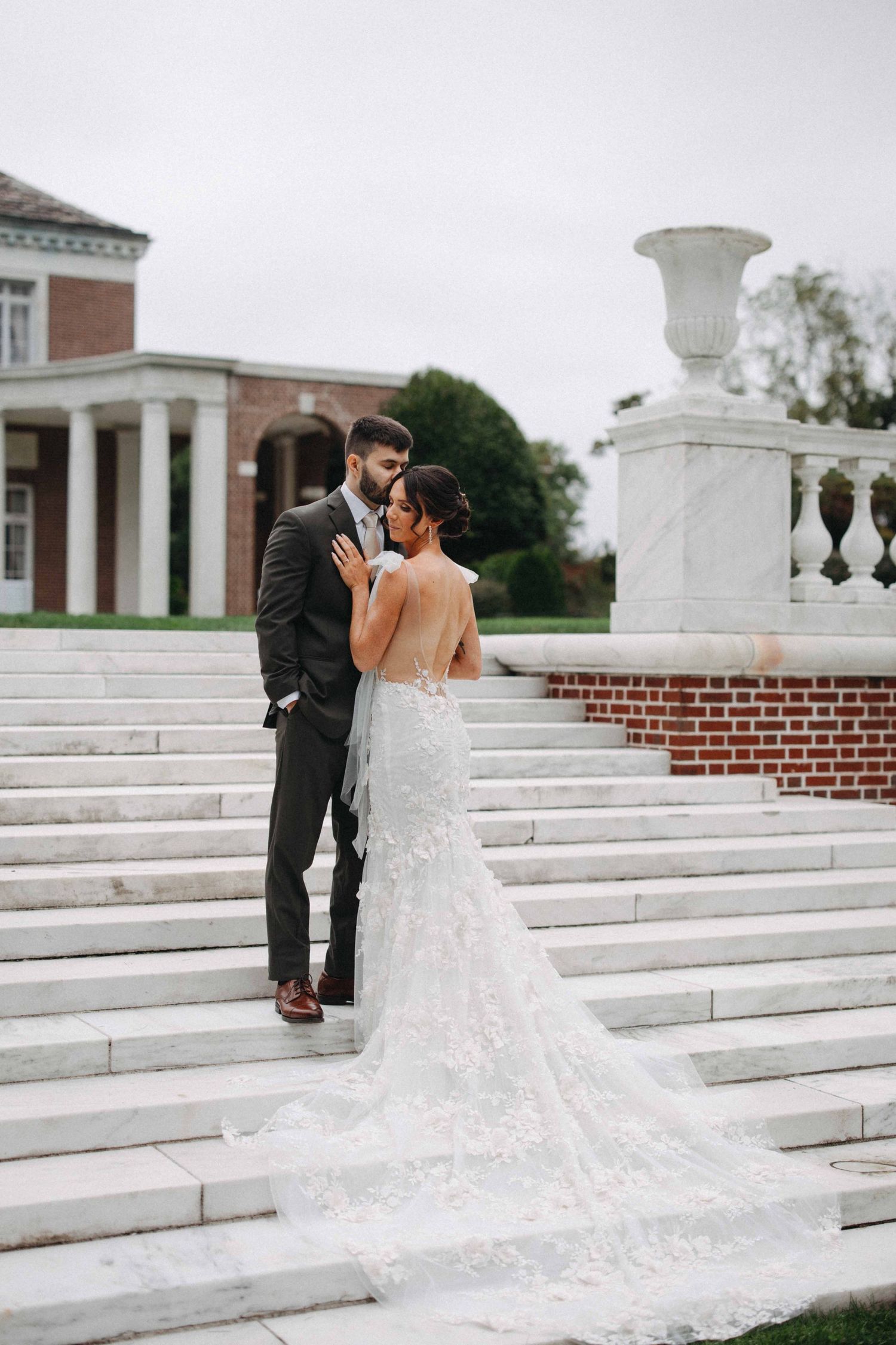 Bride in lace gown and groom in dark suit embrace on marble steps of a stately brick mansion.