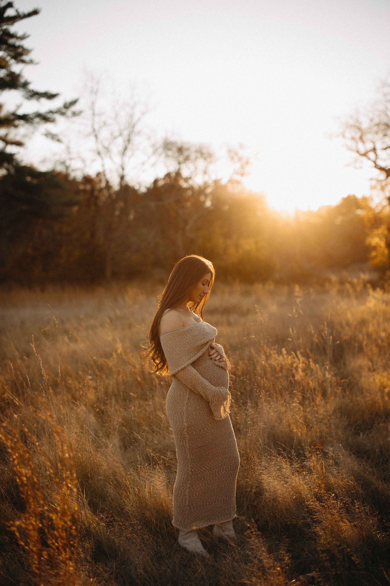 Pregnant woman in beige knit dress standing in golden field at sunset, cradling her baby bump.