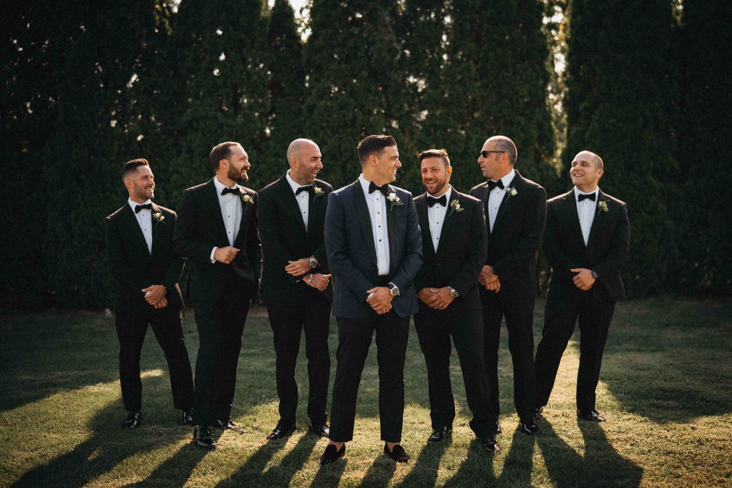 Groom and groomsmen in black tuxedos with bow ties pose together in golden sunlight outdoors.