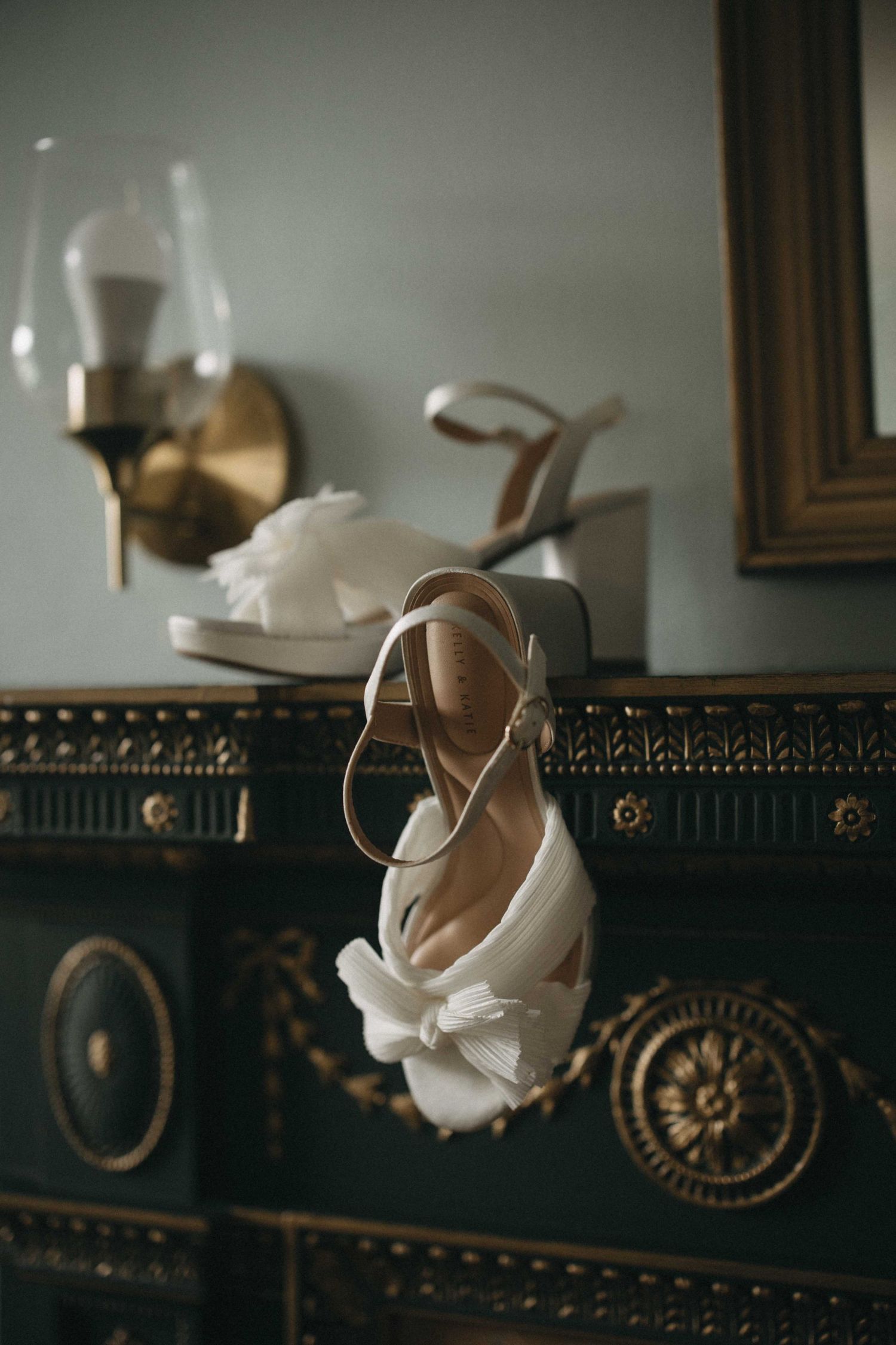 Elegant white bridal heels with floral bow detail displayed on a vintage gold-trimmed dresser in a classic dressing room.