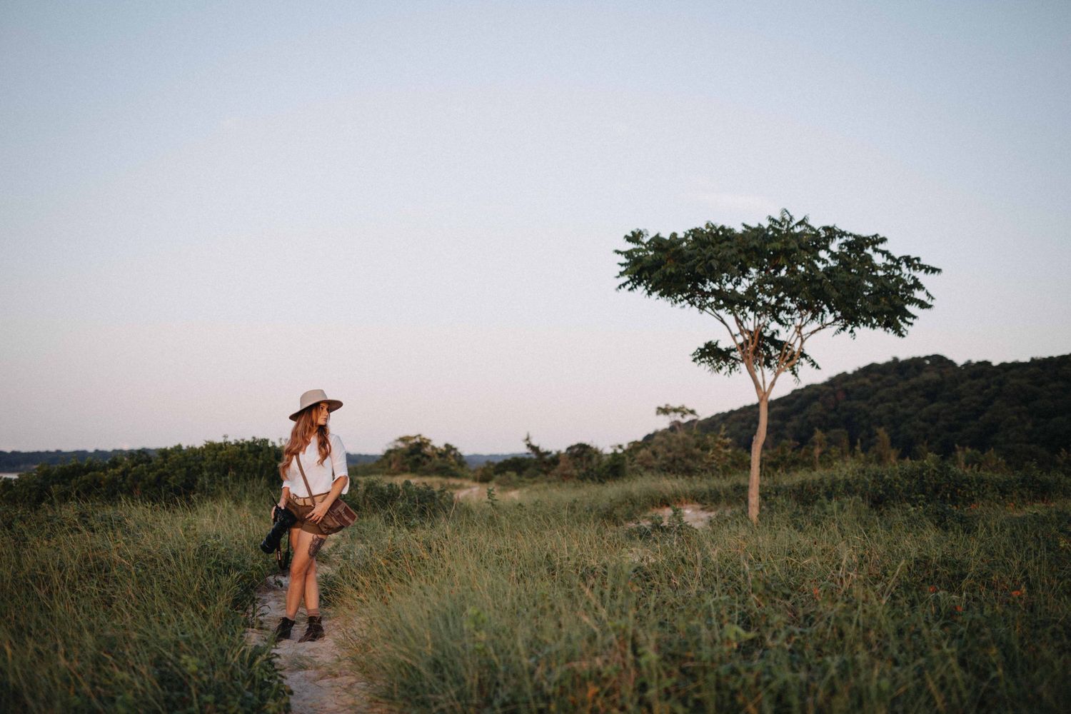 Woman in hat and white outfit walking a grassy trail near a lone tree at dusk in a scenic landscape.