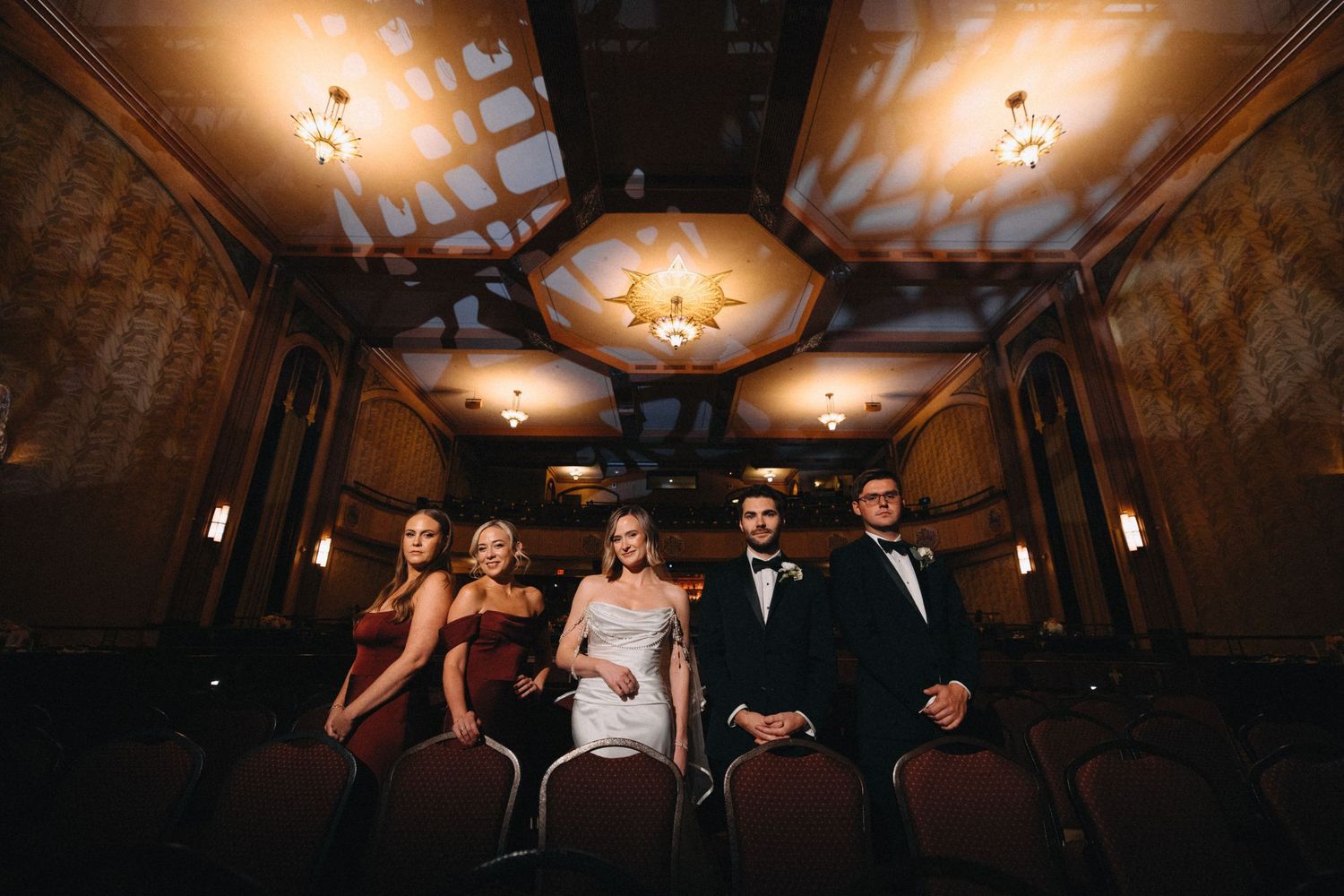 Wedding party in formal attire seated in ornate theater venue with golden ceiling and chandelier lighting.