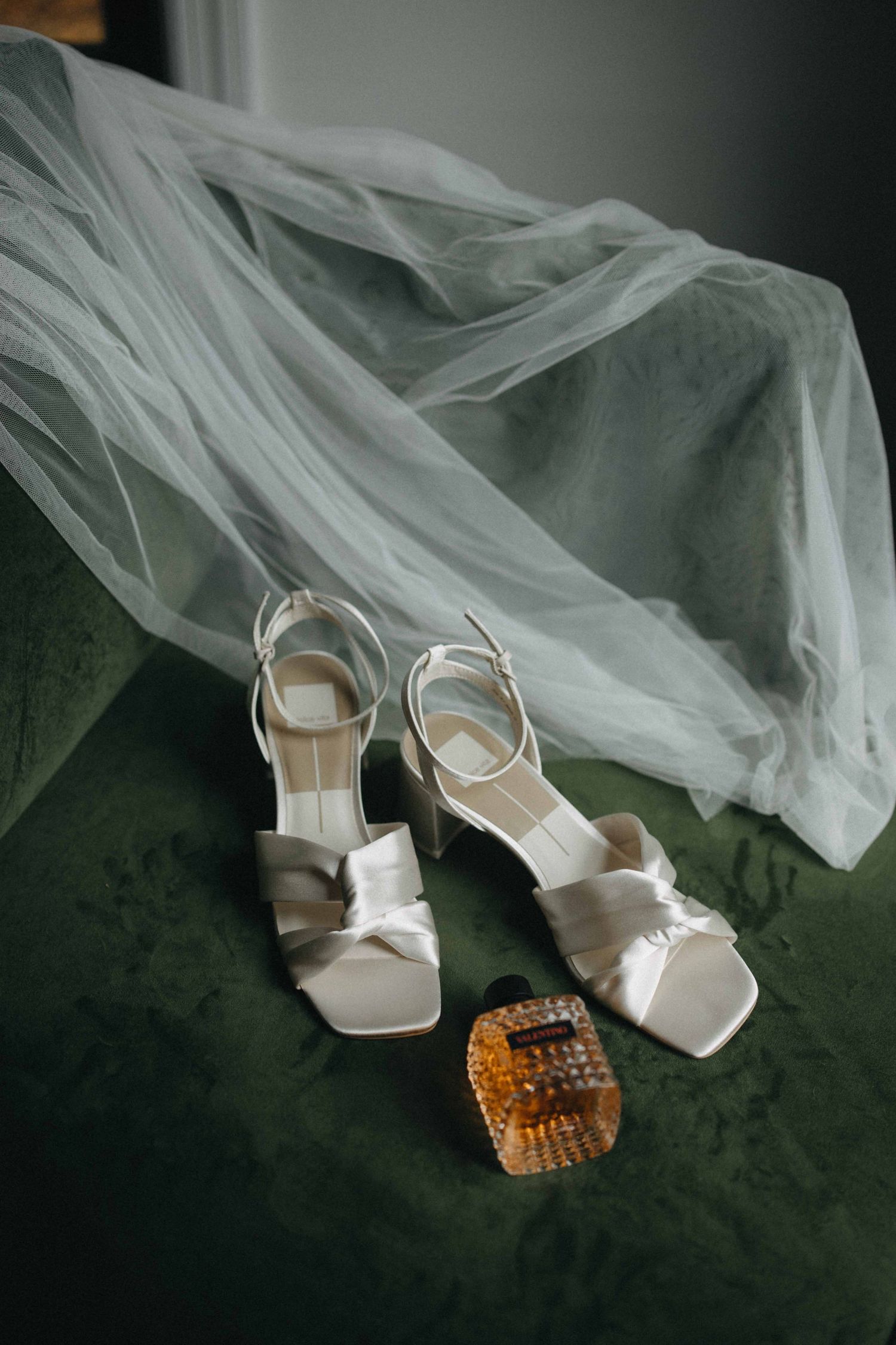 White strappy bridal heels and amber hair clip displayed on green velvet chair with tulle veil.