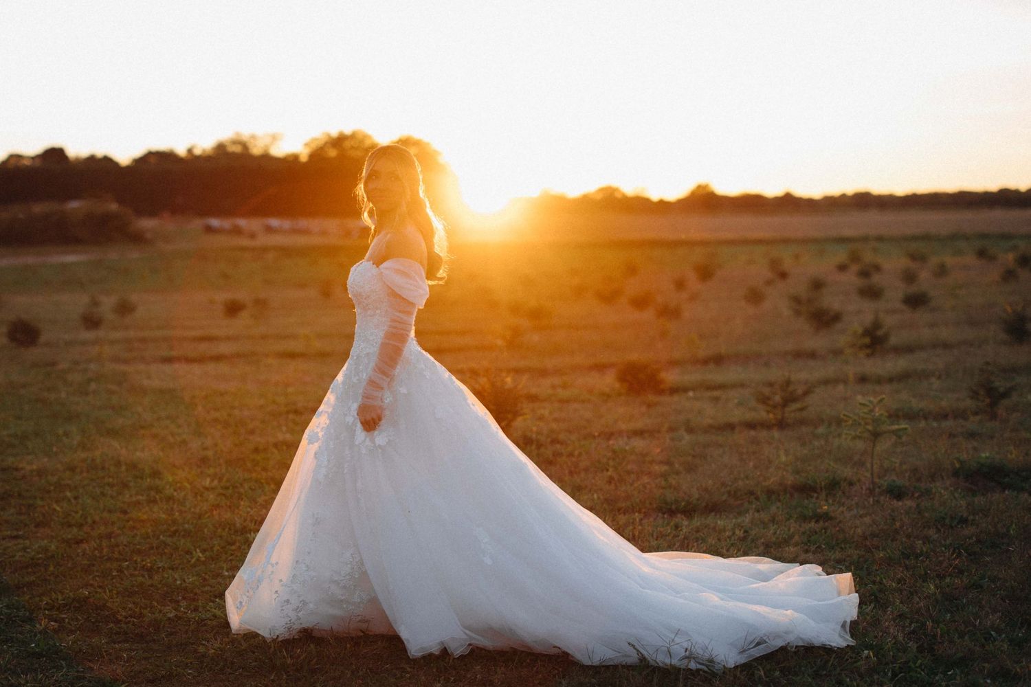 Bride in elegant off-shoulder lace wedding gown twirling in golden sunset light in open field.