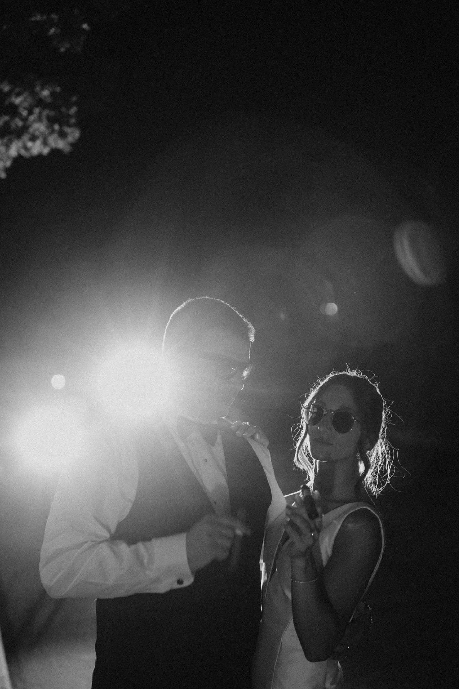 Dramatic black and white photo of bride and groom wearing sunglasses posing in front of a bright backlight at night.