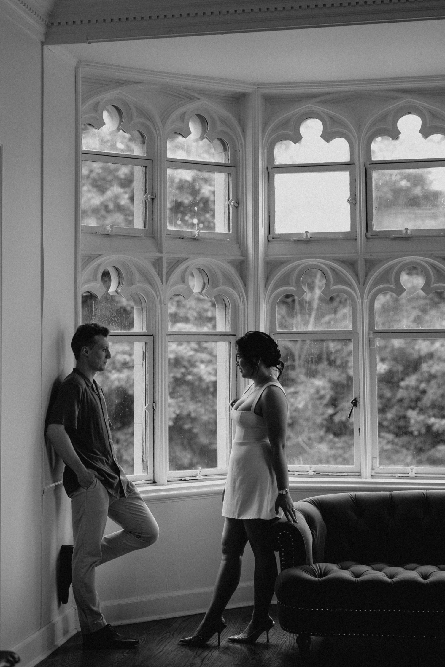 Black and white photo of a couple standing by ornate gothic arched windows in an elegant historic interior.