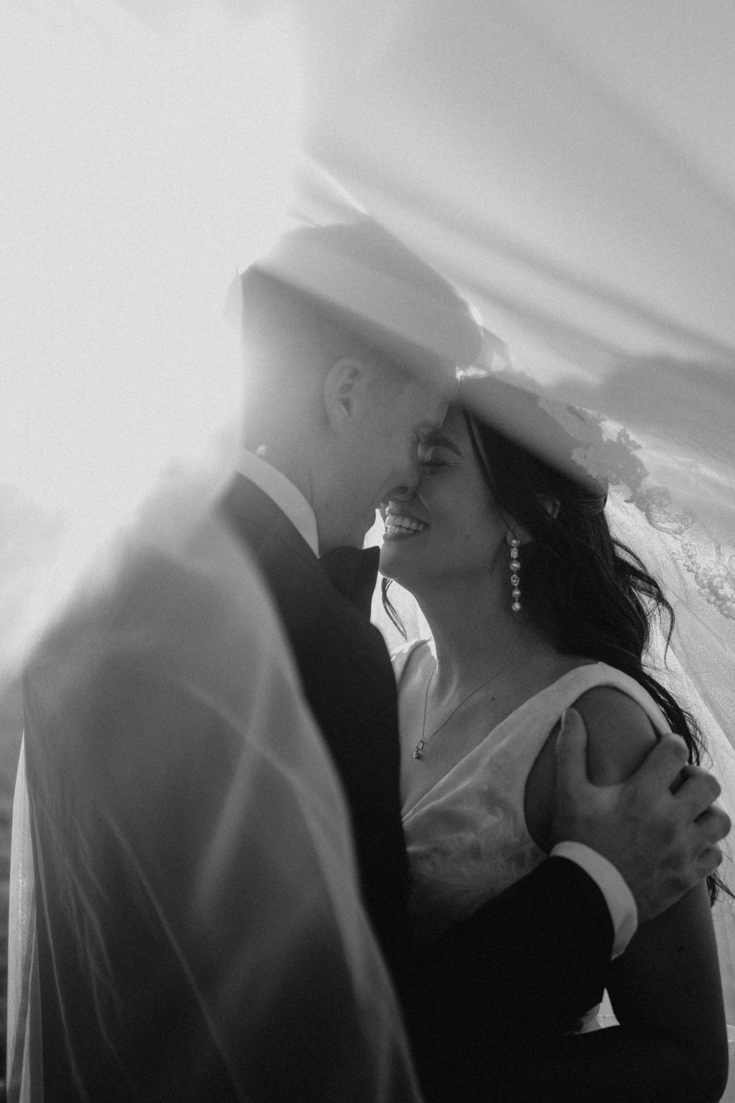 Black and white romantic portrait of bride and groom beneath flowing wedding veil.