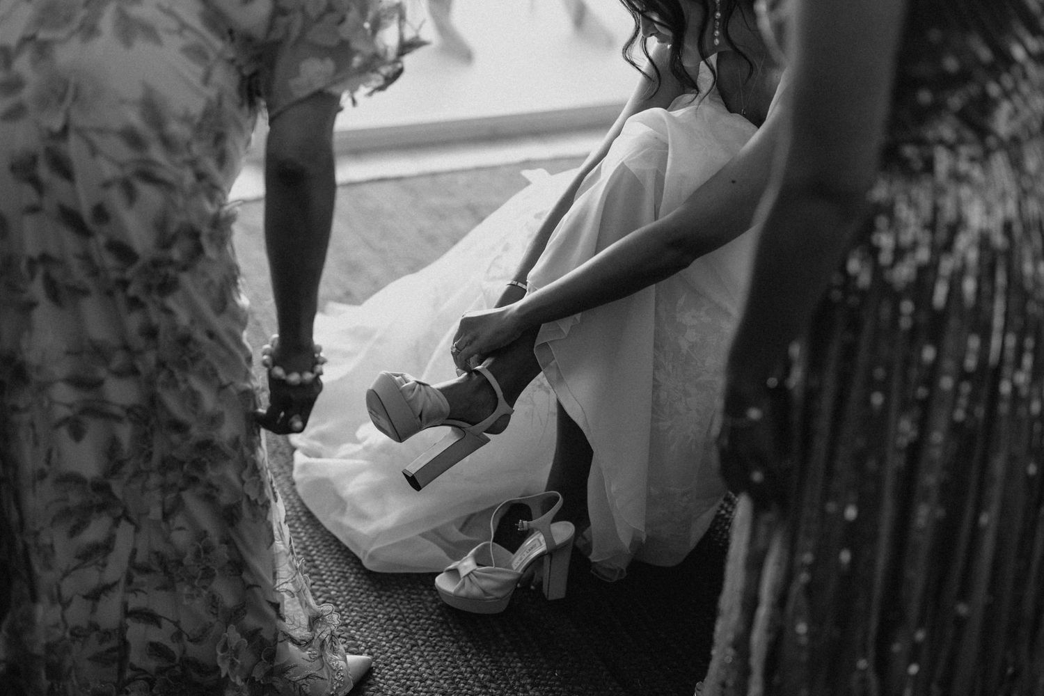 Black and white close-up of a bride being helped into her wedding heels by bridesmaids before the ceremony.