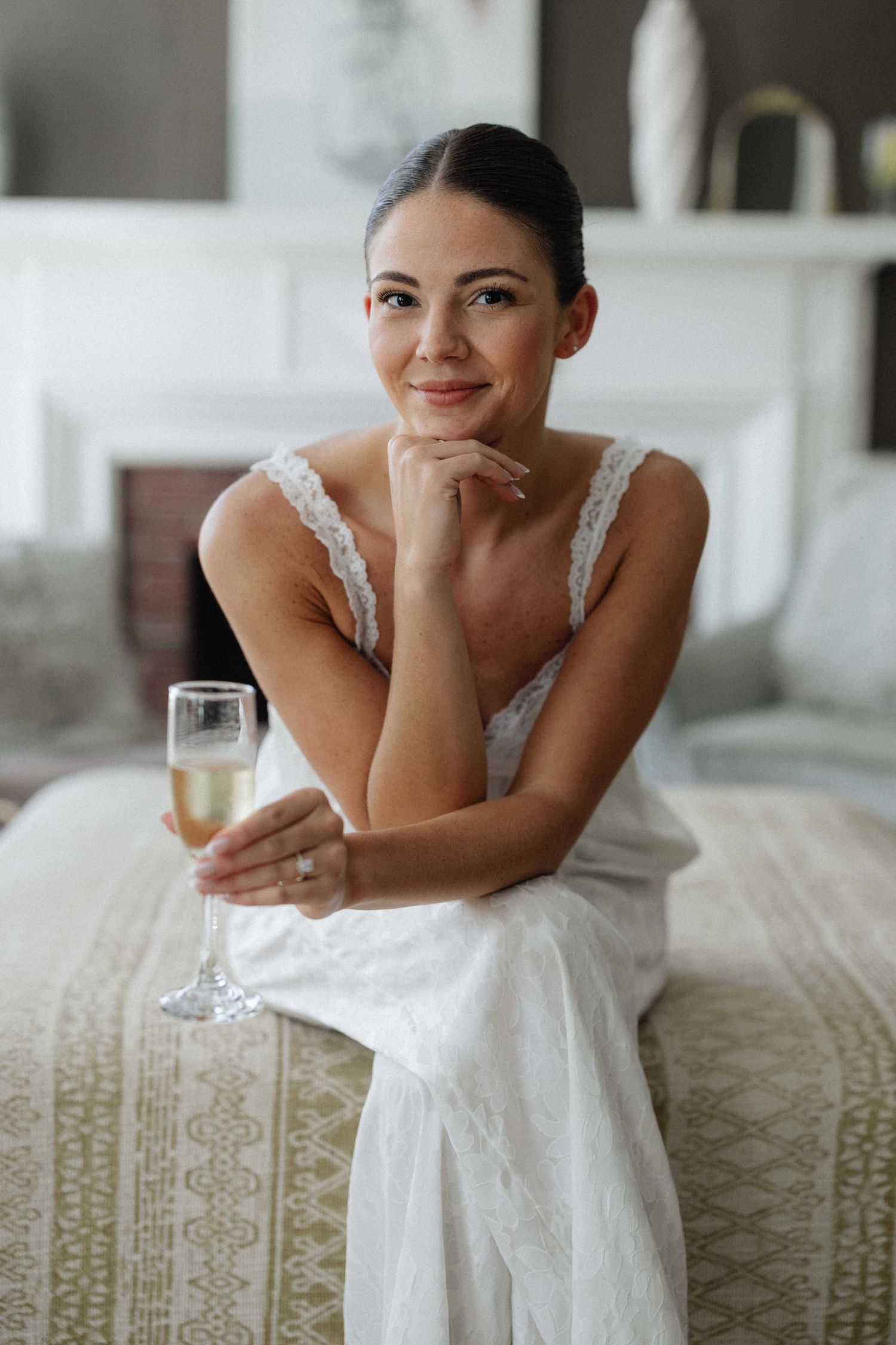 Smiling bride in white dress holding champagne glass while sitting on bed in elegant room.