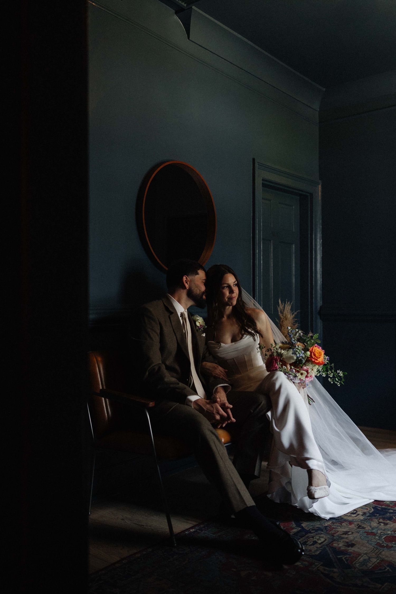 Bride and groom sit together in a moody, dark-walled room, bride holding a vibrant bouquet of orange and pink flowers.