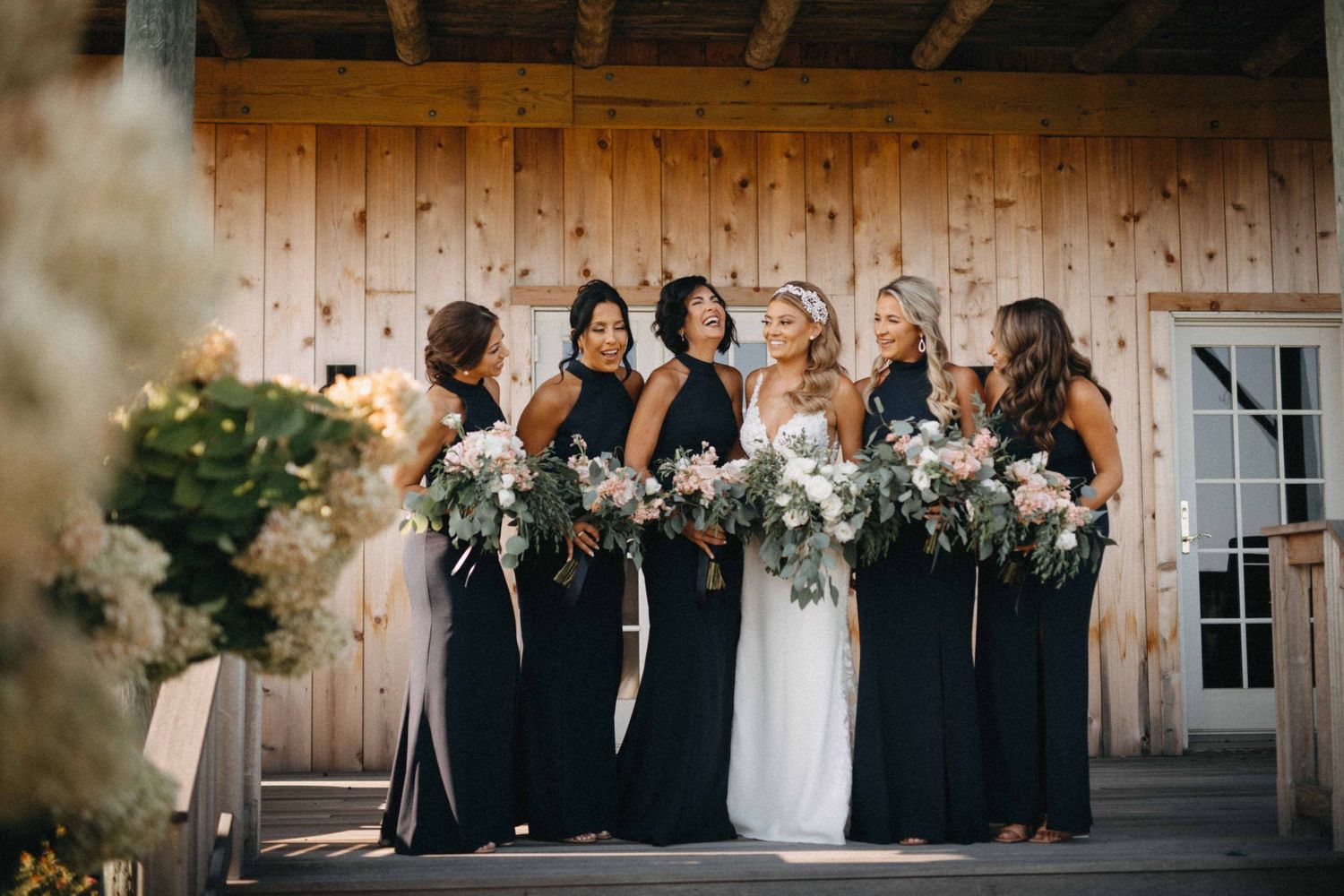 Bride and bridesmaids in black gowns holding floral bouquets laughing together outside a rustic barn.