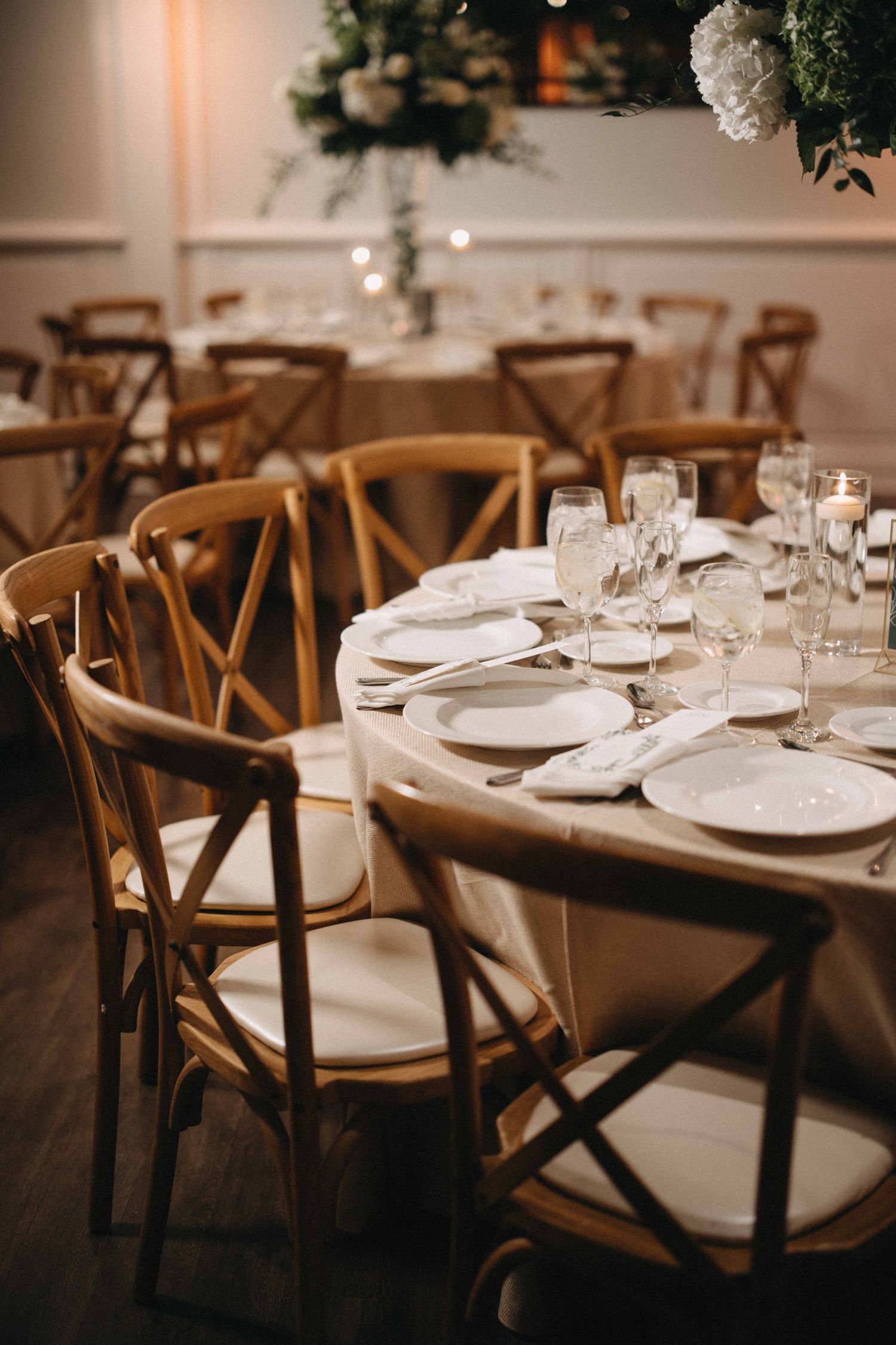 Rustic wooden cross-back chairs surrounding round tables with white linens, crystal glasses, and candles at wedding reception.