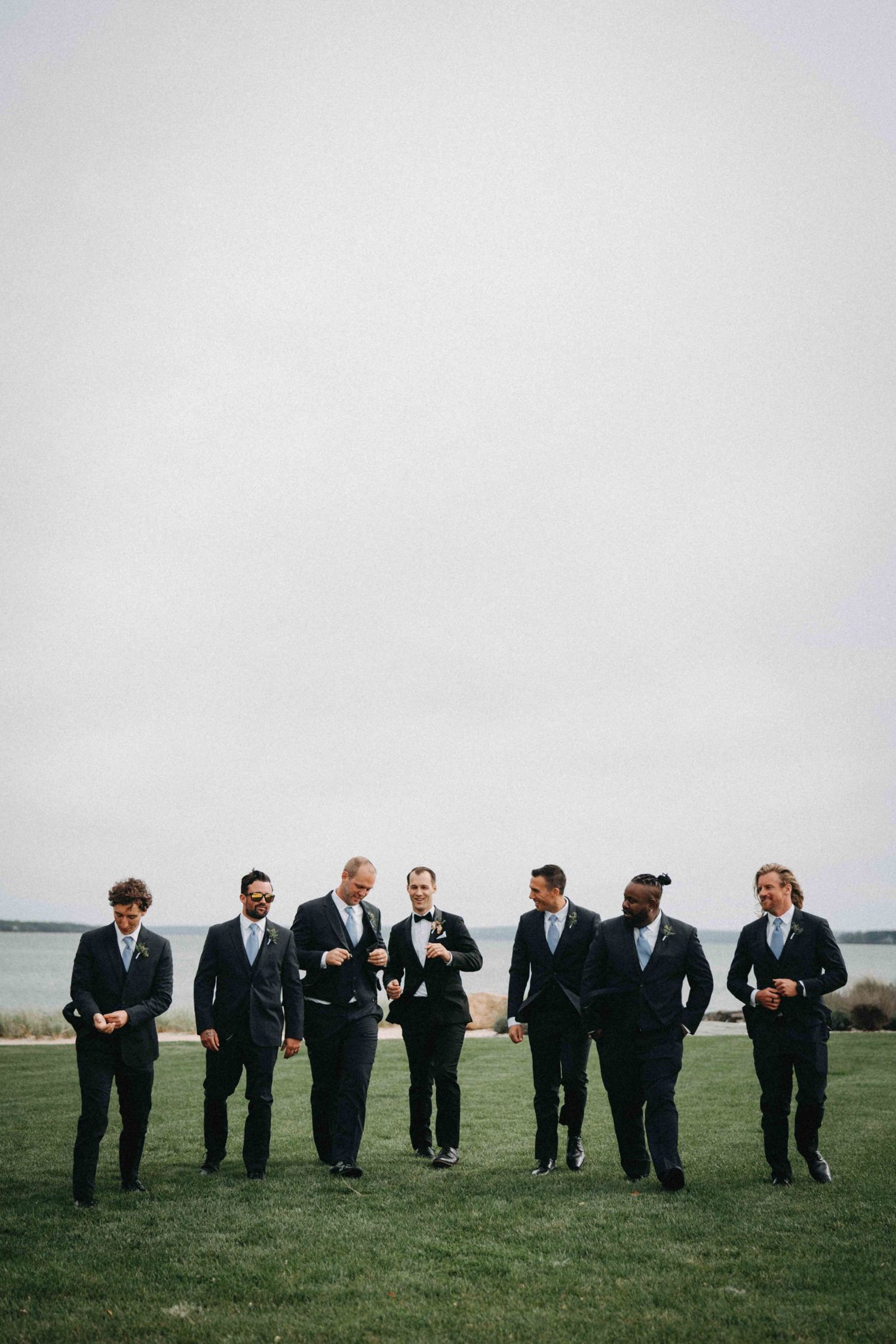 Groomsmen in dark suits walking across green lawn with water and overcast sky behind them.