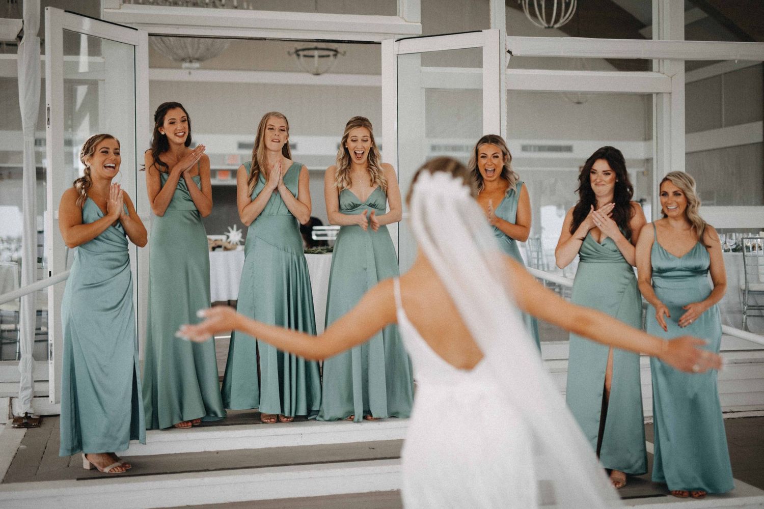 Excited bridesmaids in sage green dresses reacting to bride's first look reveal at wedding venue.