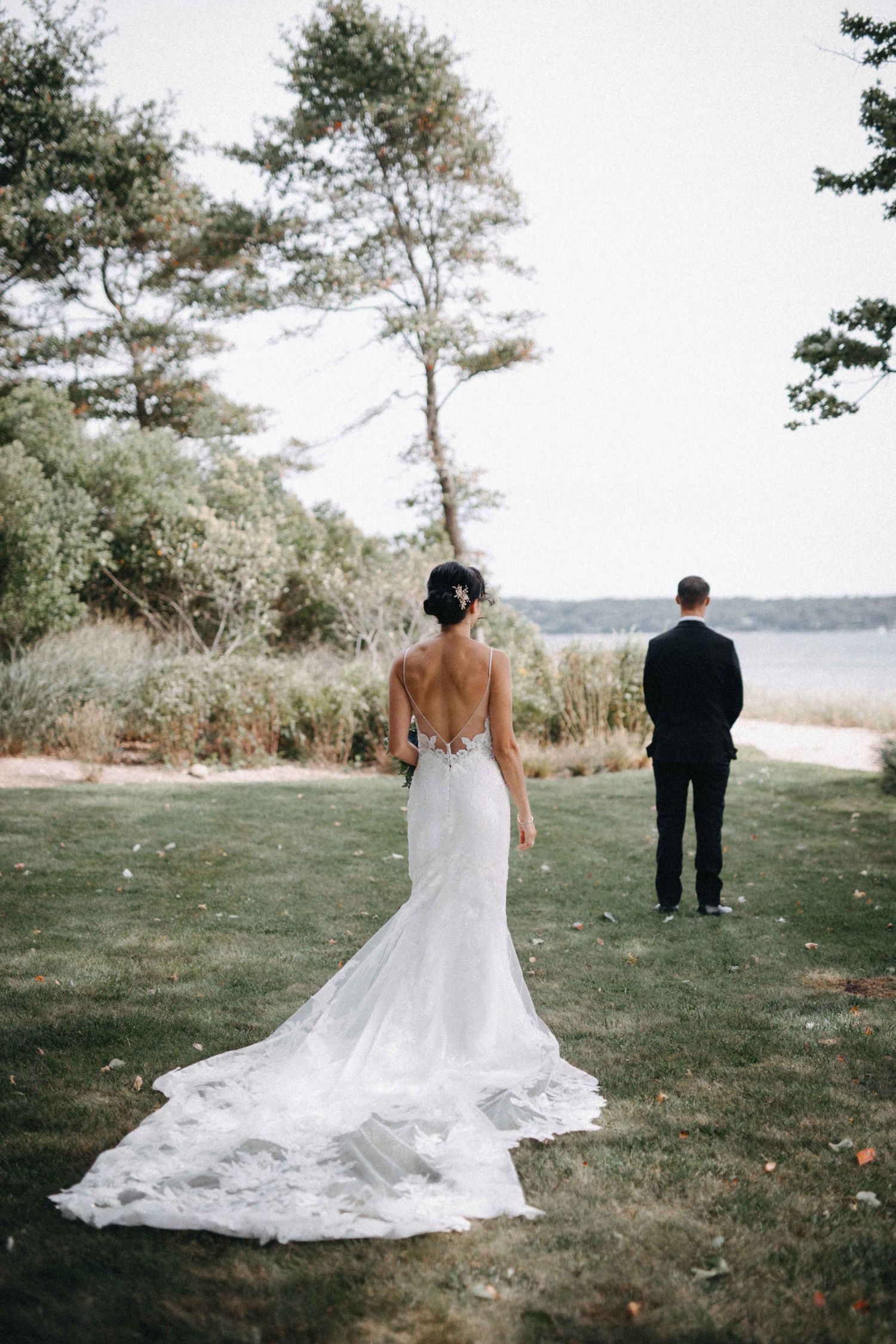 Bride in a backless lace gown with long train approaching groom near a scenic waterfront landscape.