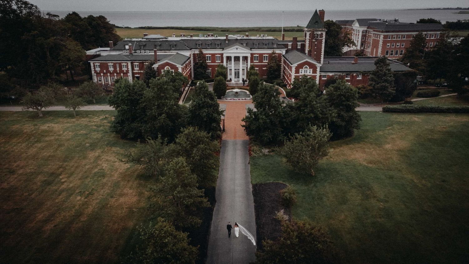 Aerial drone view of a grand brick colonial wedding venue surrounded by lush green trees near the waterfront.