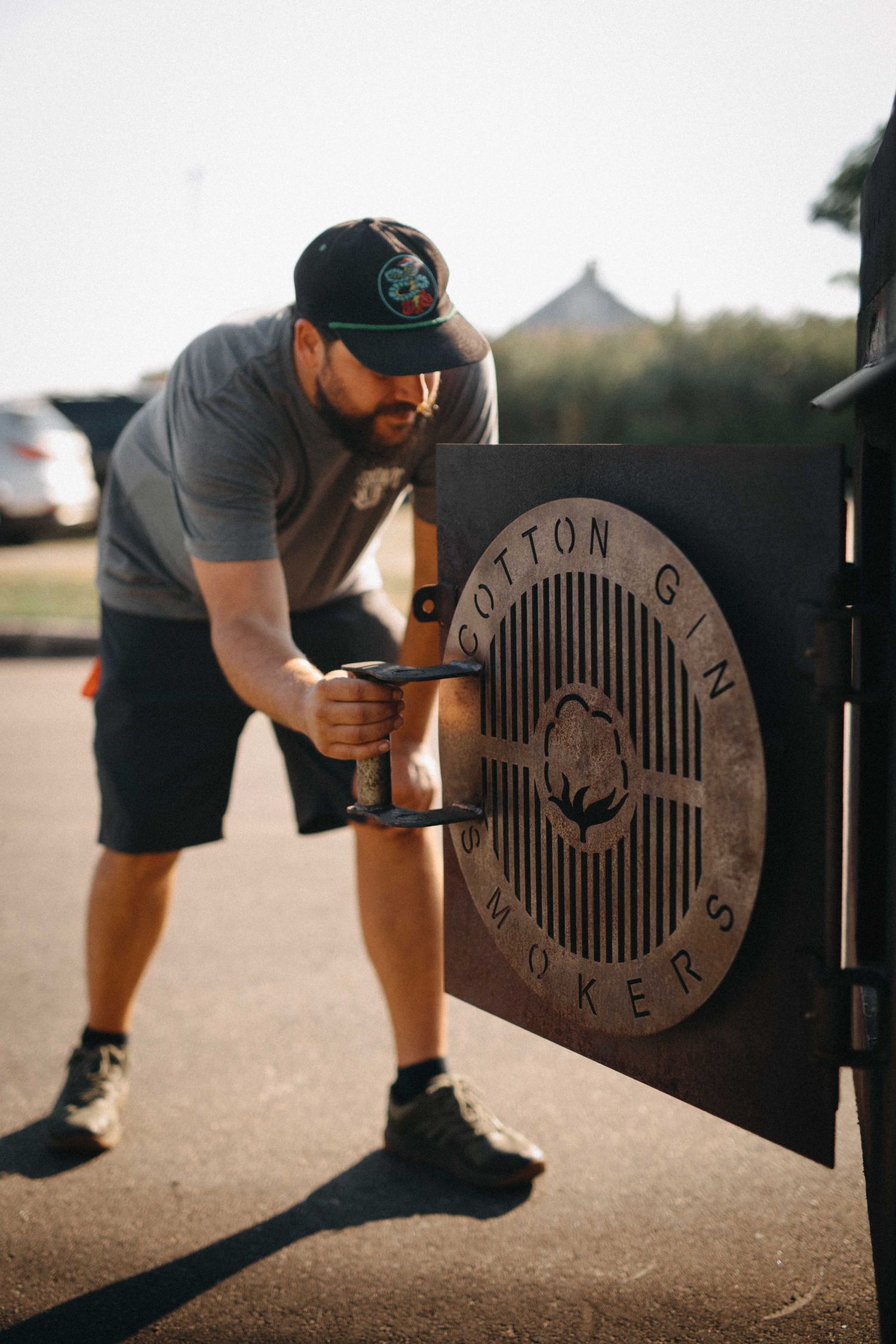 Bearded man opening a Cotton Gin Smokers branded BBQ smoker door outdoors on a sunny day.