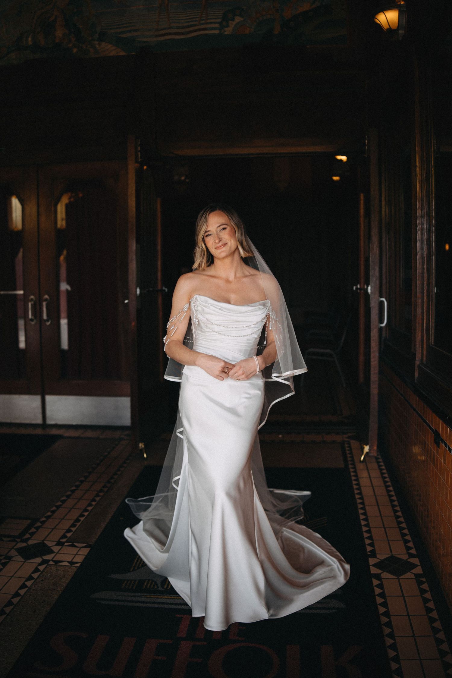 Bride in strapless white satin gown with veil poses in elegant dark wood venue corridor.