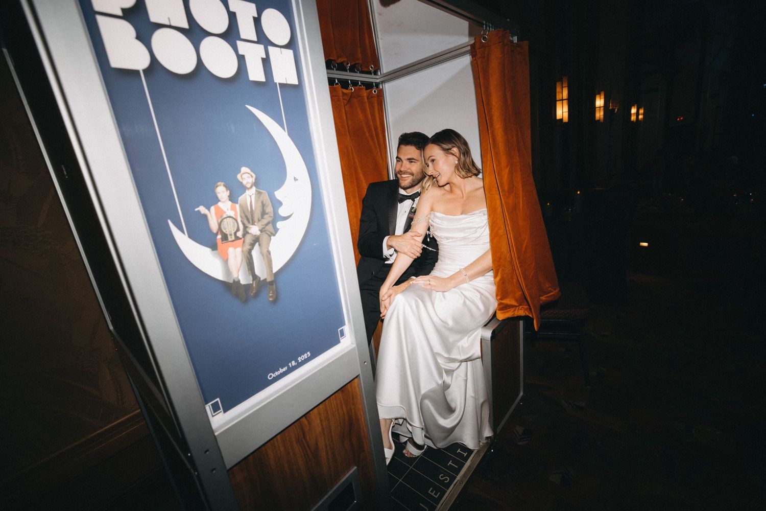 Newlywed couple poses together outside a classic photo booth at their elegant wedding reception.