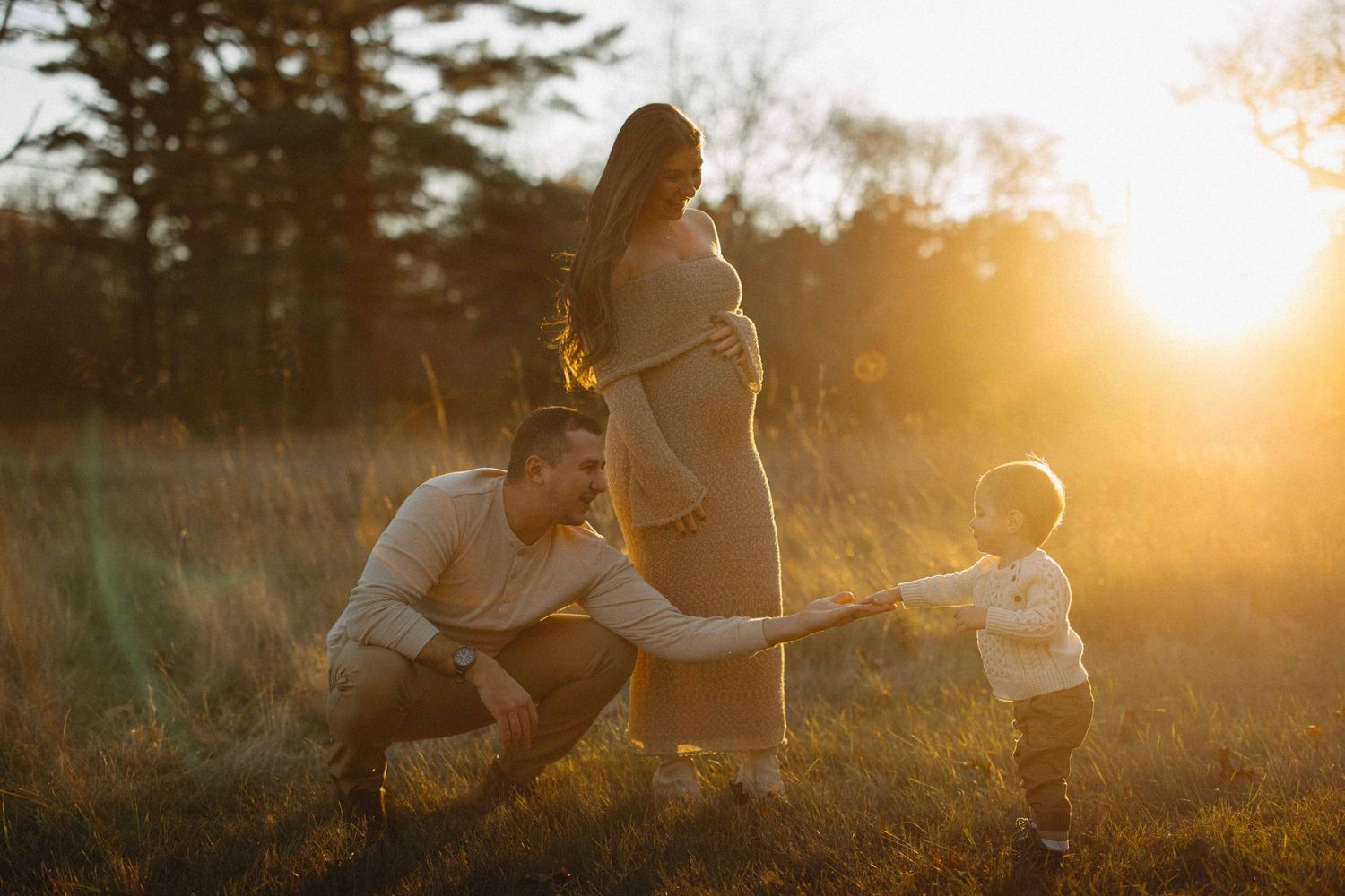 Pregnant woman and husband interact with toddler in golden sunset light in a field during maternity photoshoot.