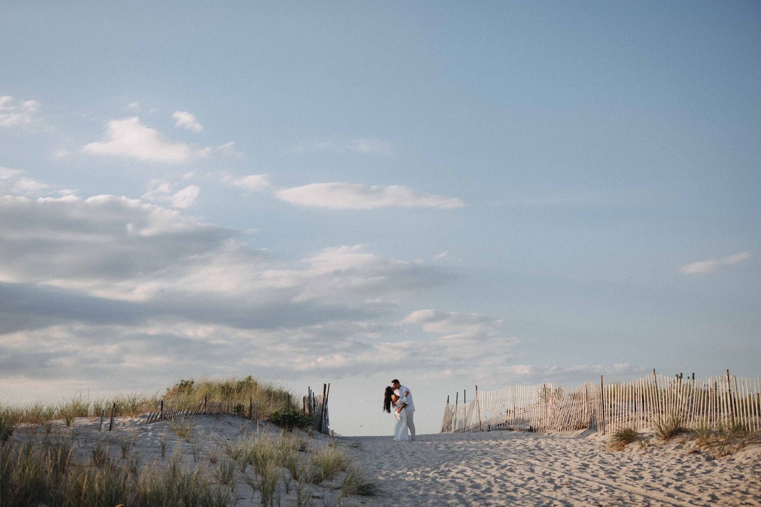 Couple sharing a kiss on a sandy beach pathway surrounded by dunes under a blue sky.