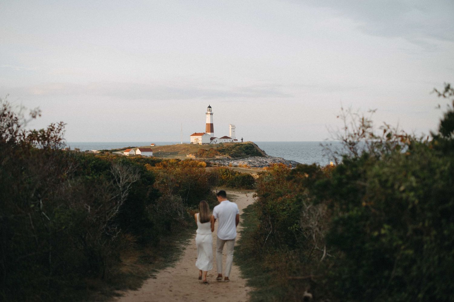 Couple strolling along a dirt trail toward a scenic oceanside lighthouse.