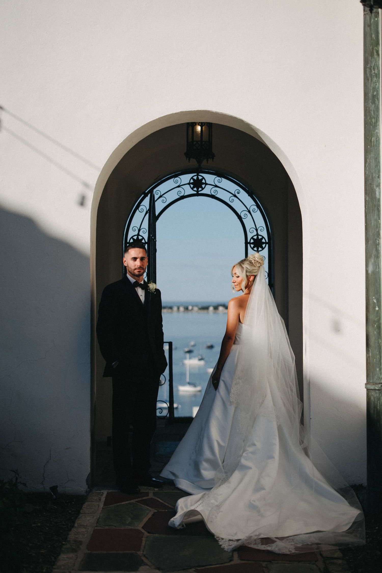 Bride and groom standing in ornate arched doorway with ocean view beyond.