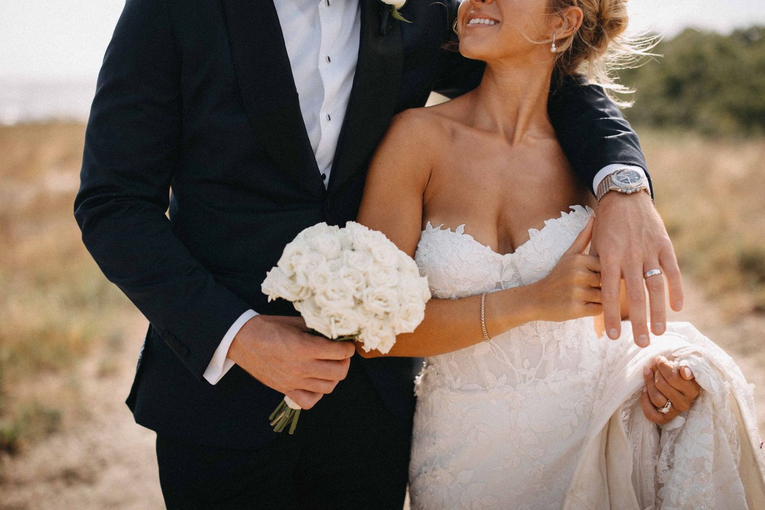 Bride and groom embrace outdoors, groom holding white bouquet while bride wears strapless lace gown.