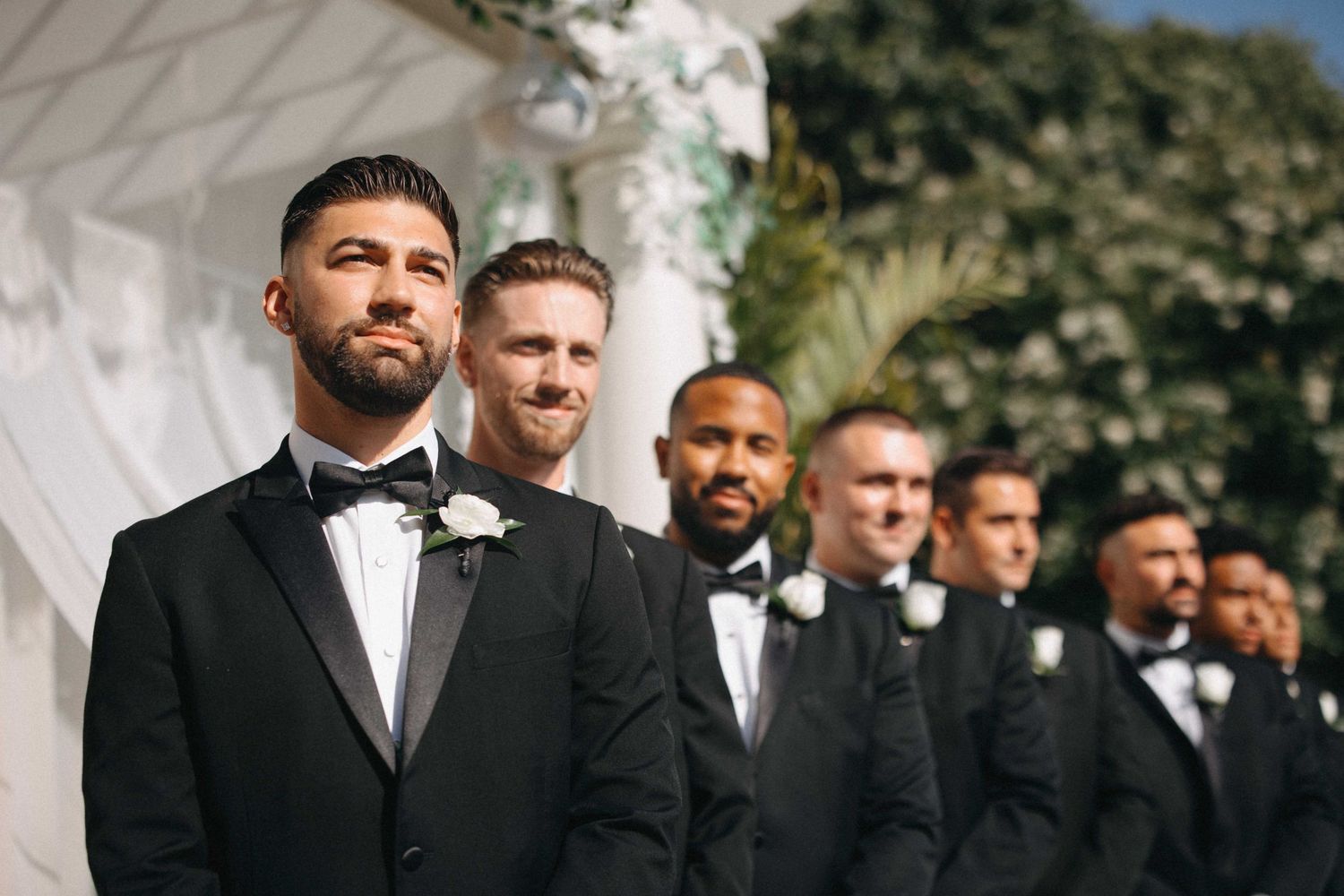 Dapper groom and groomsmen in black tuxedos lined up at an outdoor wedding ceremony altar.
