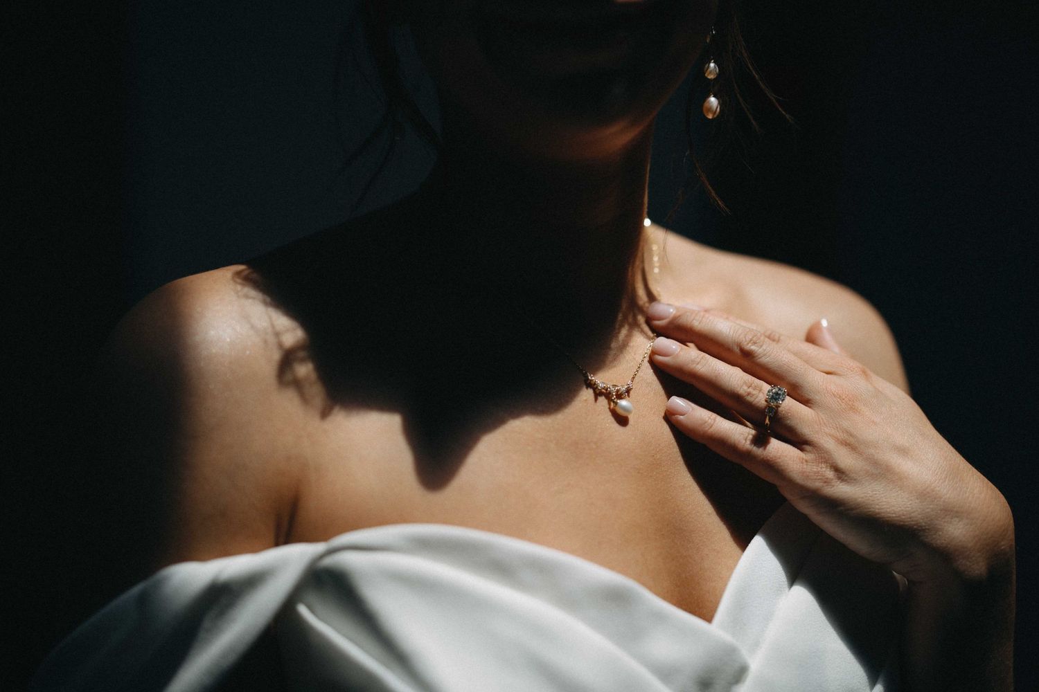 Close-up of a bride in off-shoulder white dress touching a delicate gold necklace in dramatic shadow lighting.