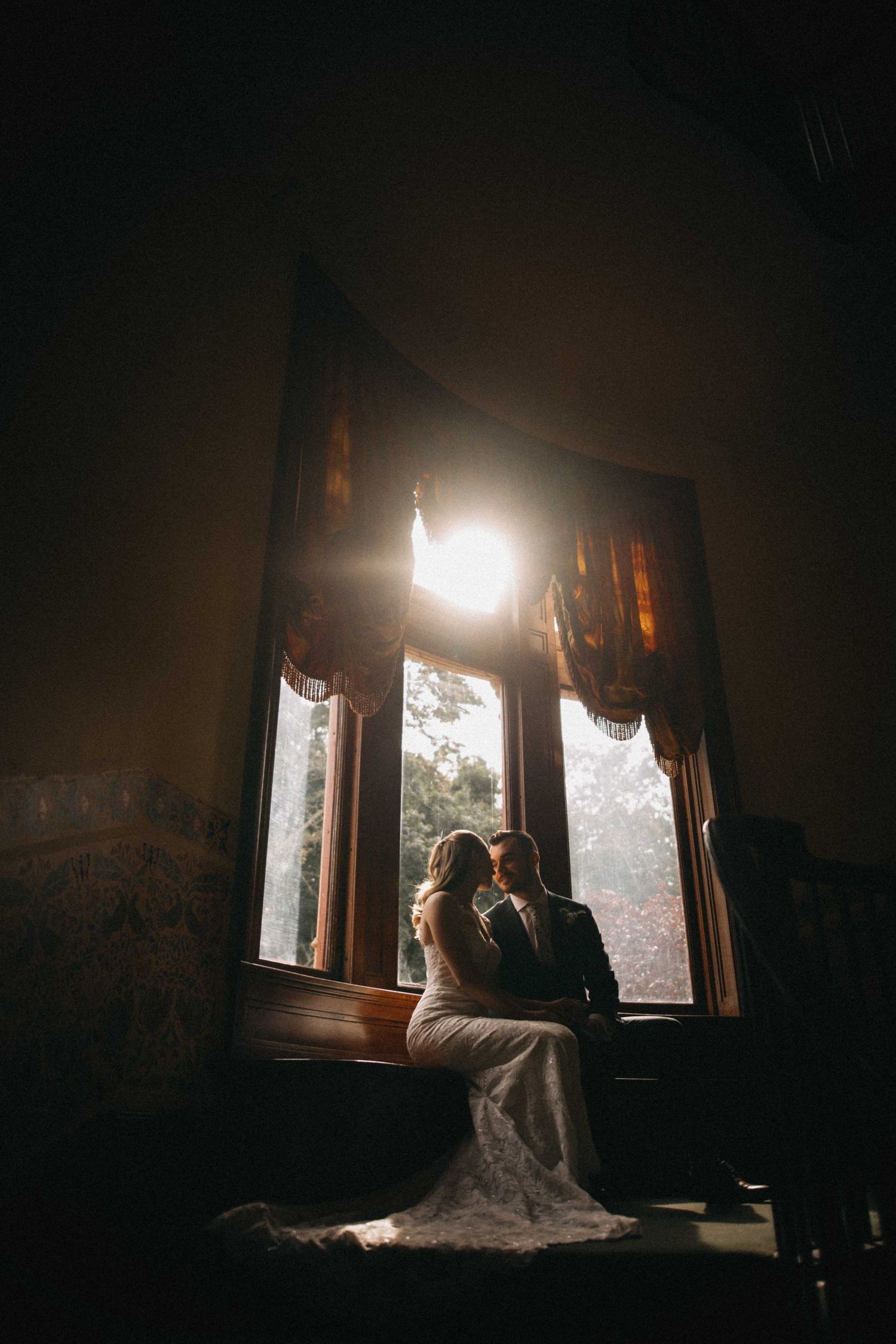 Bride and groom silhouetted against a sunlit window in a dark elegant room during their wedding portrait session.