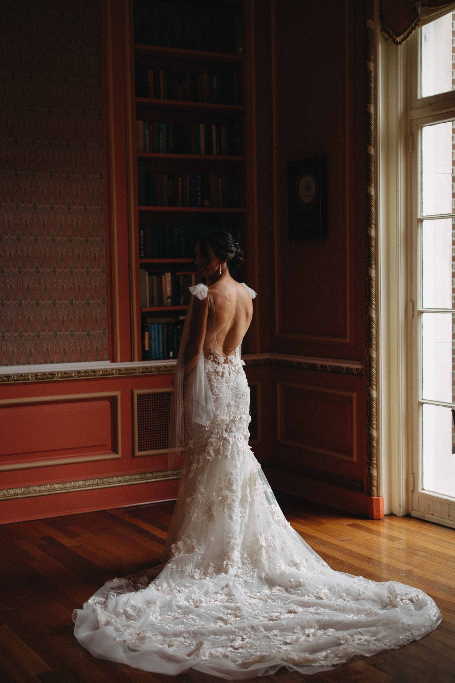 Bride in a backless lace wedding gown with floral detail standing by a window in an ornate library room.