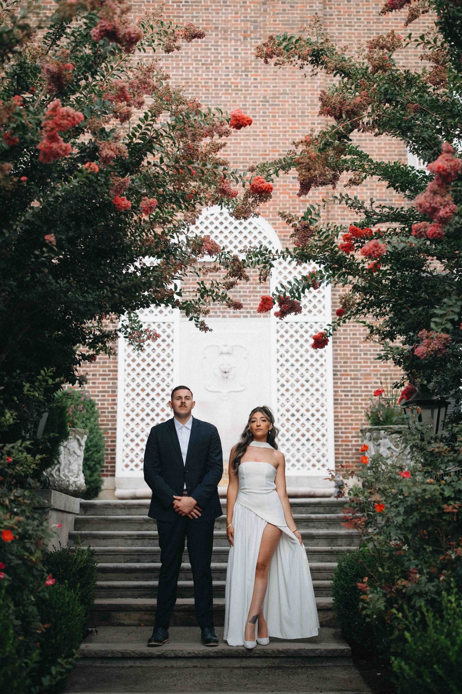 Couple standing beneath a floral archway of red blooms against a brick wall.