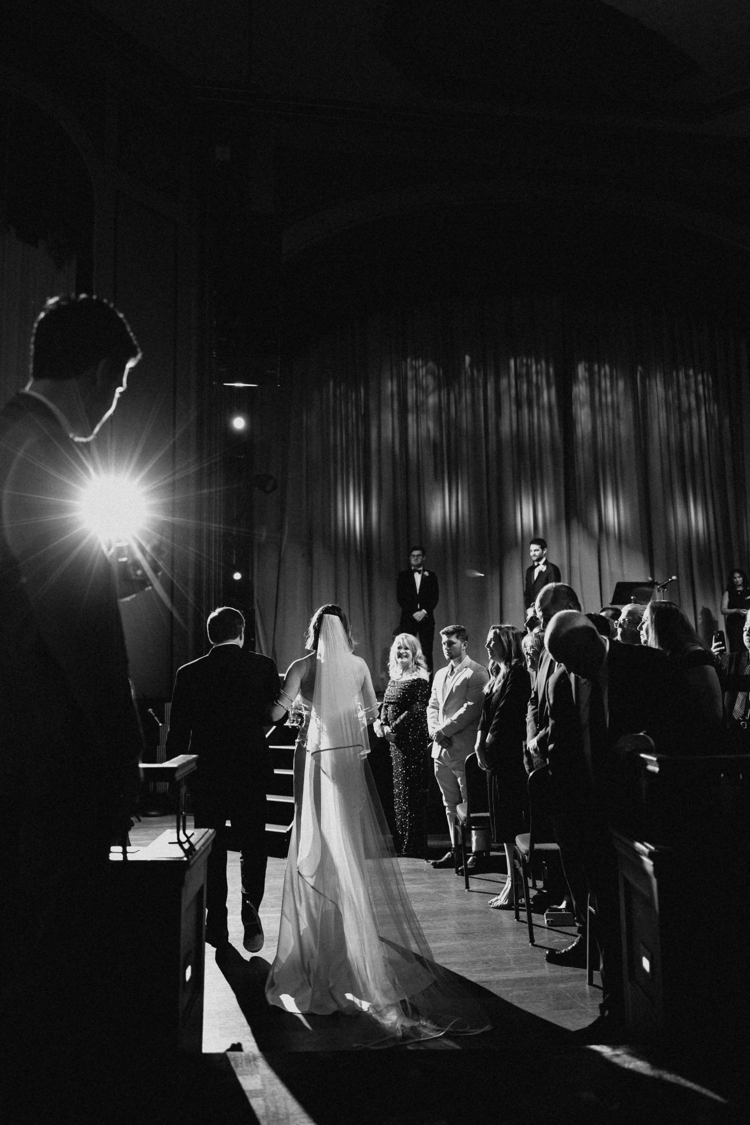 Black and white photo of bride walking down aisle in veil toward altar in dramatic lit venue.