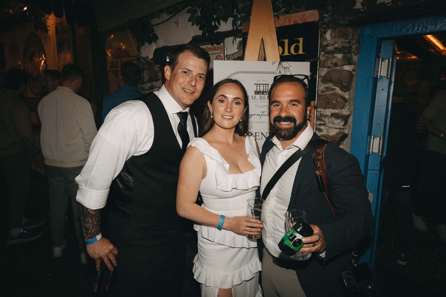 Two men and a woman smiling together at a lively wedding reception venue at night.